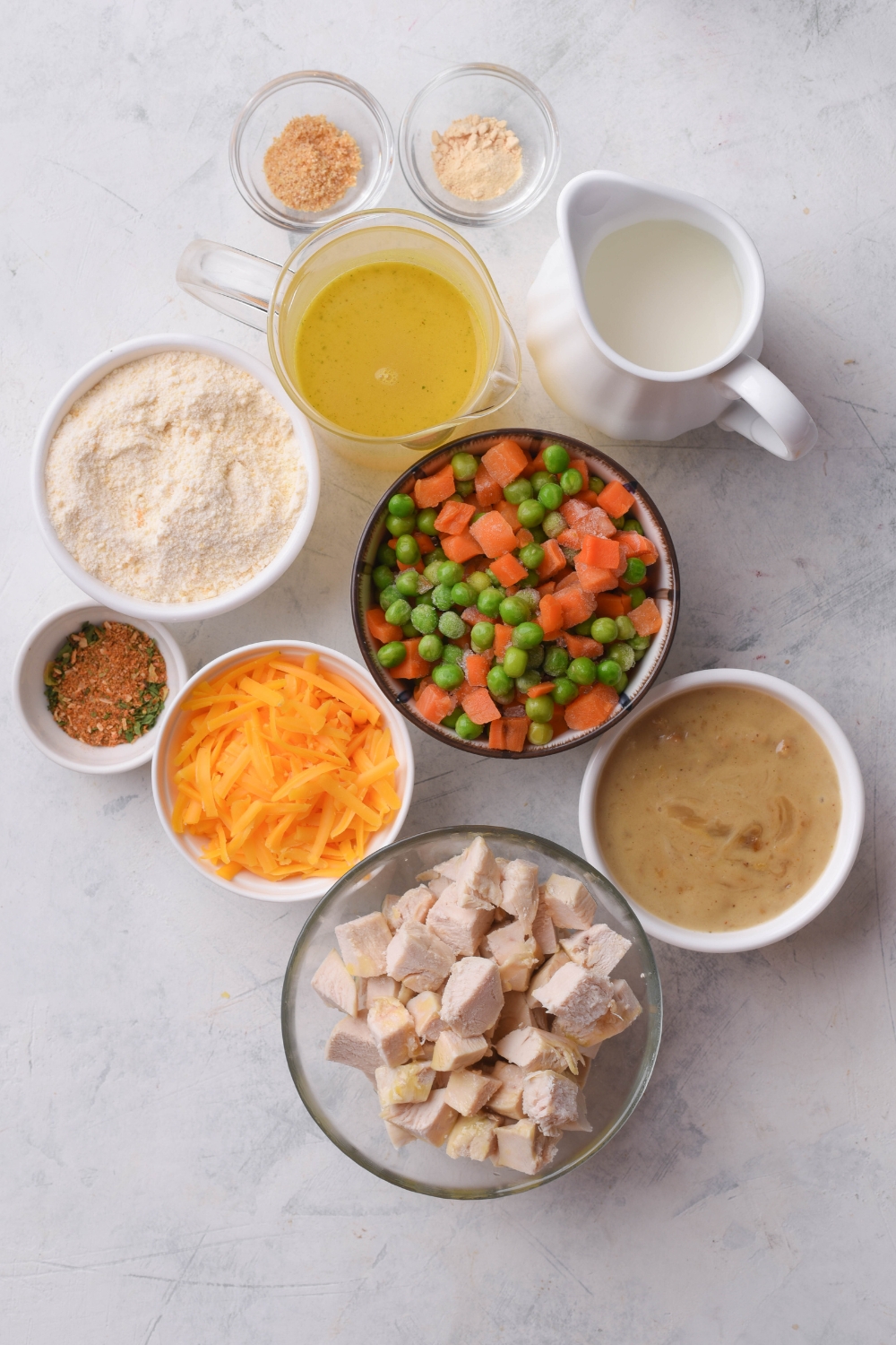 An assortment of ingredients including bowls of peas and carrots, diced chicken, condensed soup, broth, cream, shredded cheese, a flour mixture, and several small bowls of seasonings all on a grey counter.