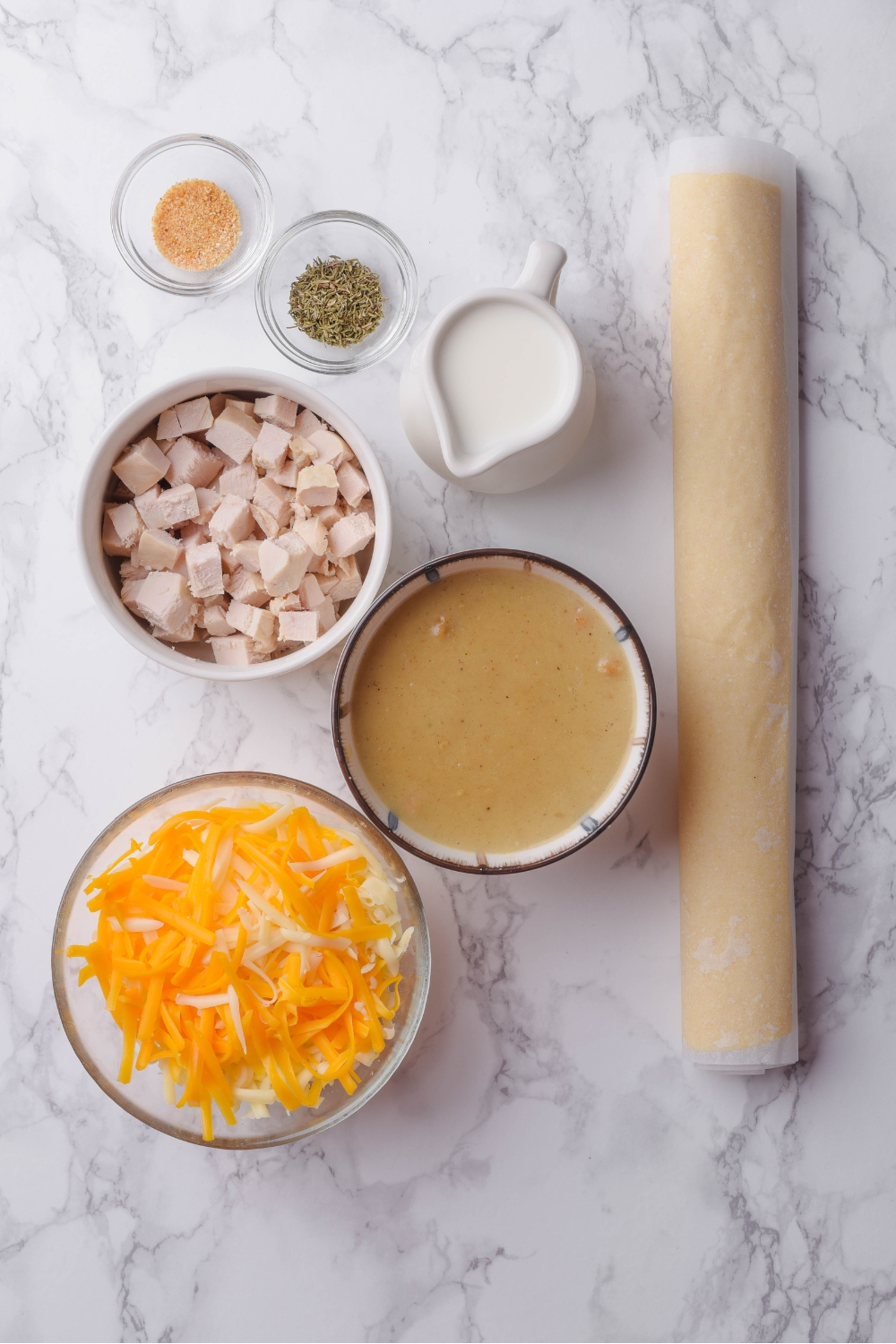 An assortment of ingredients including a roll of crescent roll dough and bowls of condensed soup, milk, shredded cheese, diced chicken, and spices.