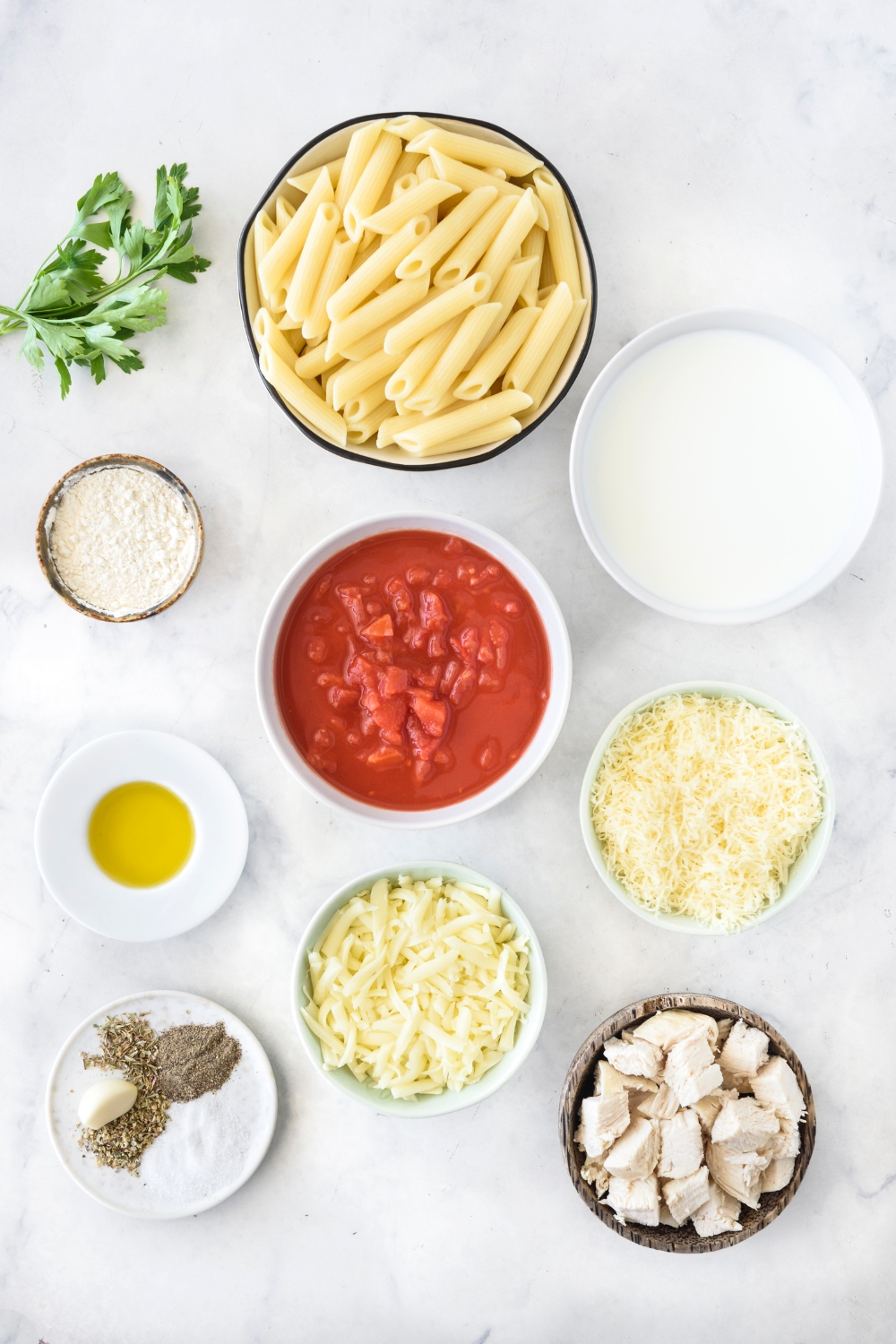 An assortment of ingredients including bowls of dried pasta, red sauce, shredded cheese, diced chicken, oil, and milk.
