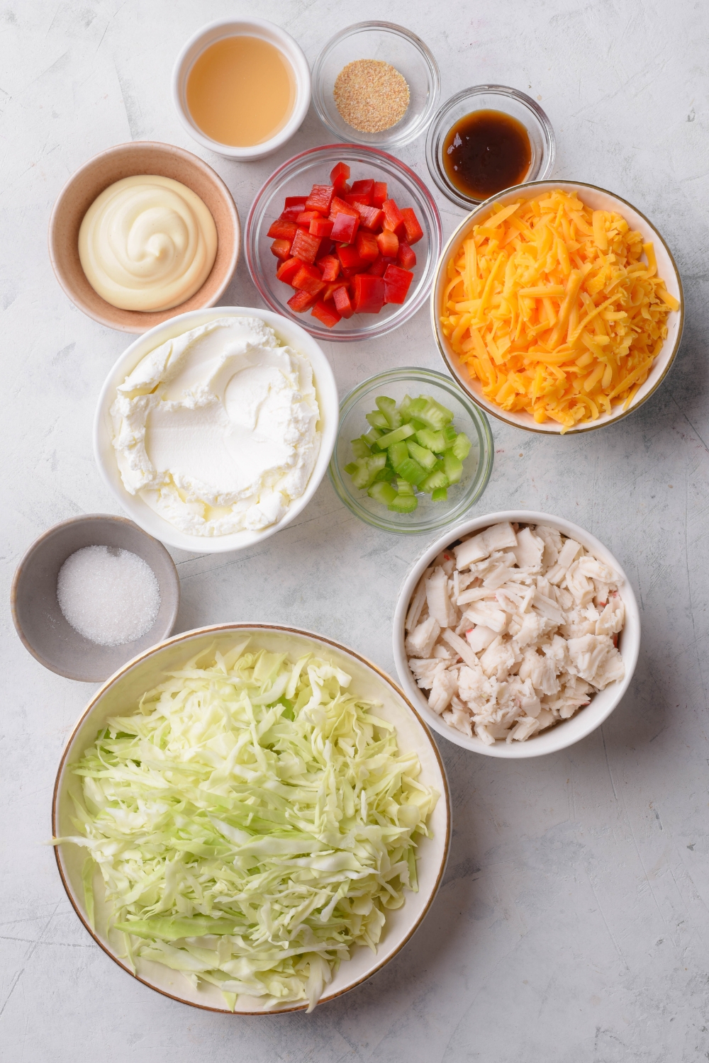 An assortment of ingredients including bowls of shredded cheese, cream cheese, sliced cabbage, crab meat, soy sauce, diced red peppers, diced celery, mayonnaise, and seasonings, on a grey counter.