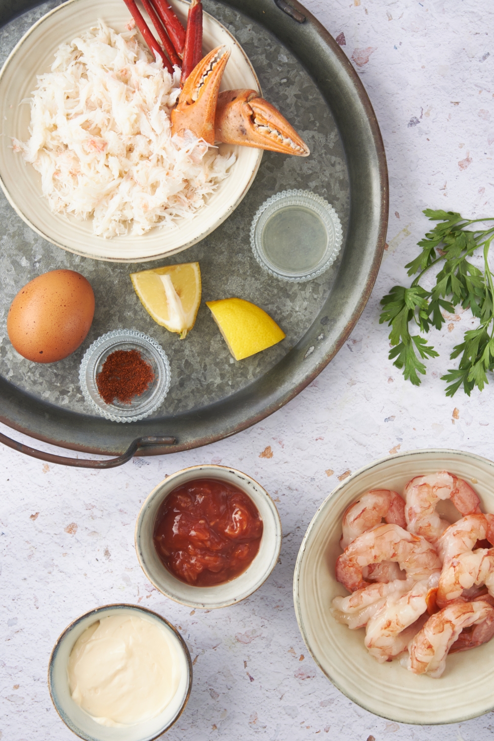 An assortment of ingredients including bowls of raw shrimp, cocktail sauce, mayonnaise, crab meat, spices, a couple of lemon wedges, and an egg.