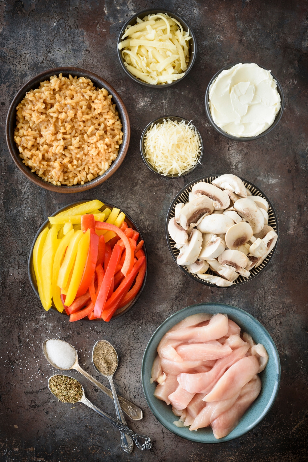 An assortment of ingredients including bowls of raw chicken strips, mushrooms, red and yellow peppers, rice, shredded cheese, cream cheese, and spoonfuls of spices.