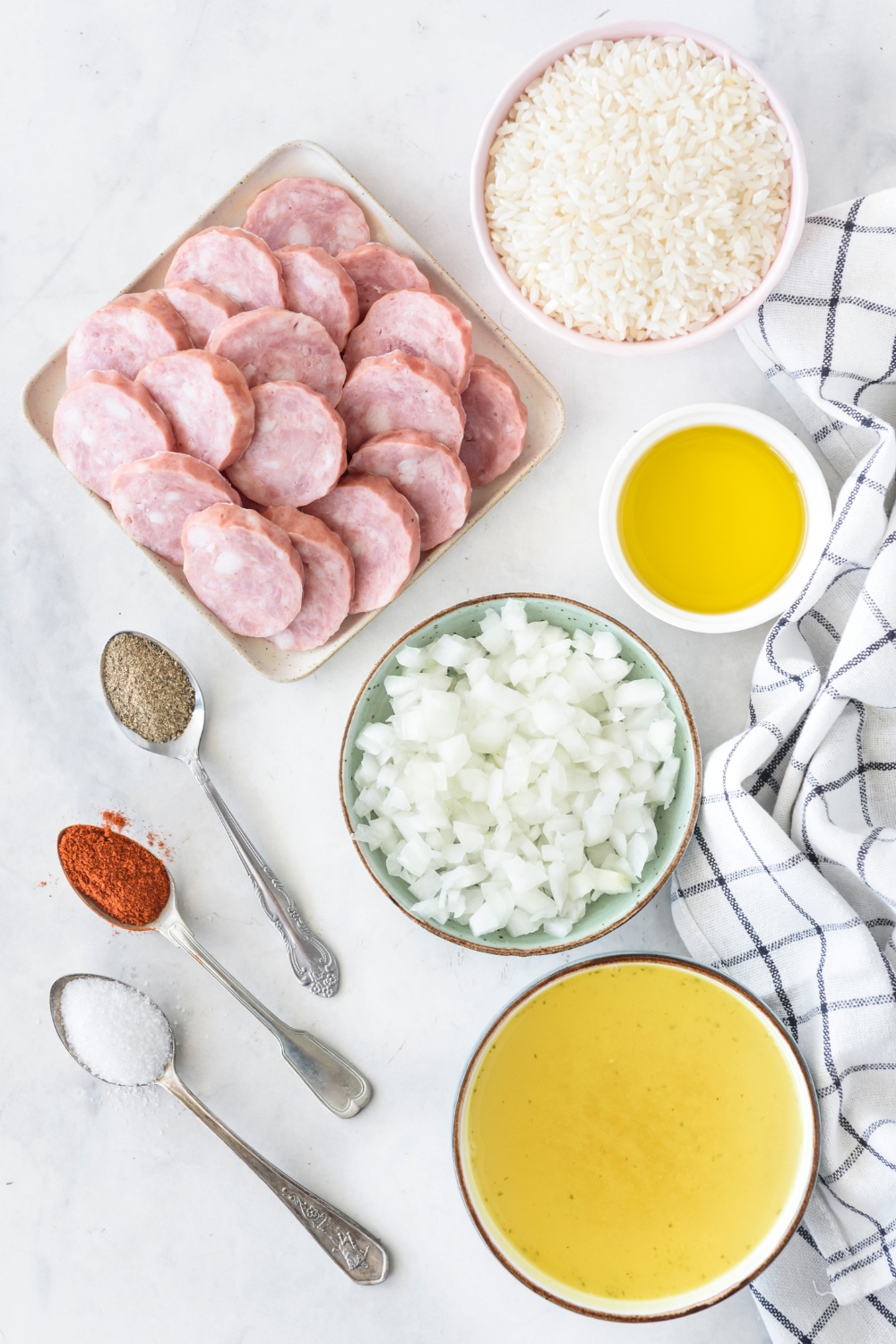 An assortment of ingredients including a plate of sliced sausages and bowls of rice, broth, diced onion, oil, and three spoons holding spices.