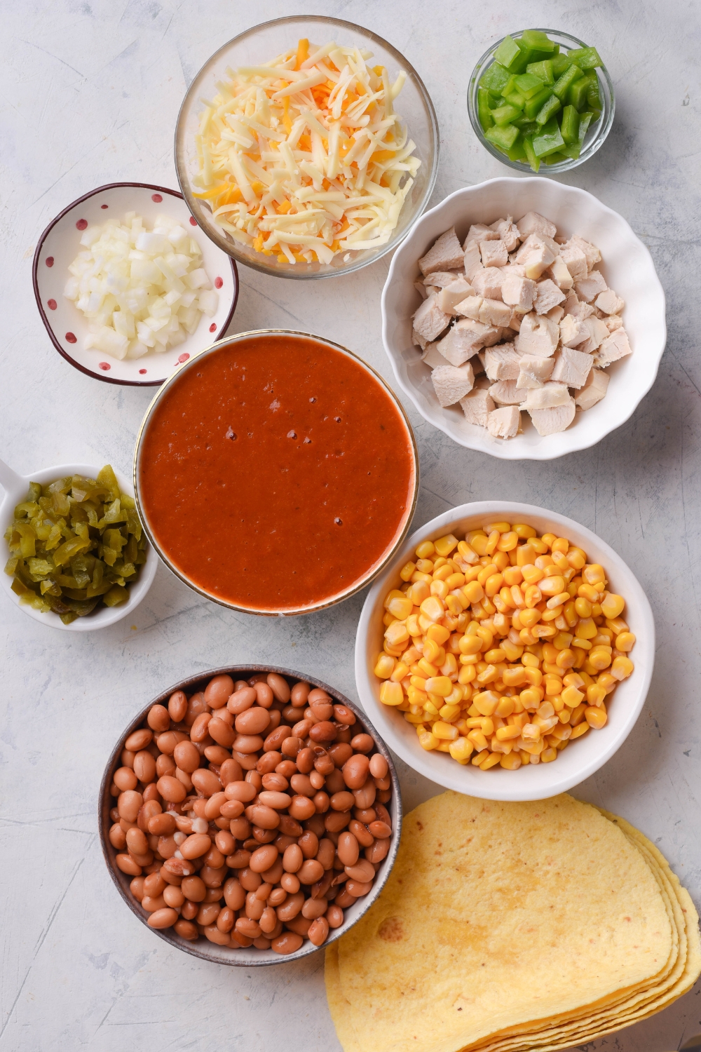 An assortment of ingredients including bowls of enchilada sauce, diced chicken, pinto beans, corn, shredded cheese, diced green peppers, diced onion, green chiles, and a stack of tortillas all on a white counter.
