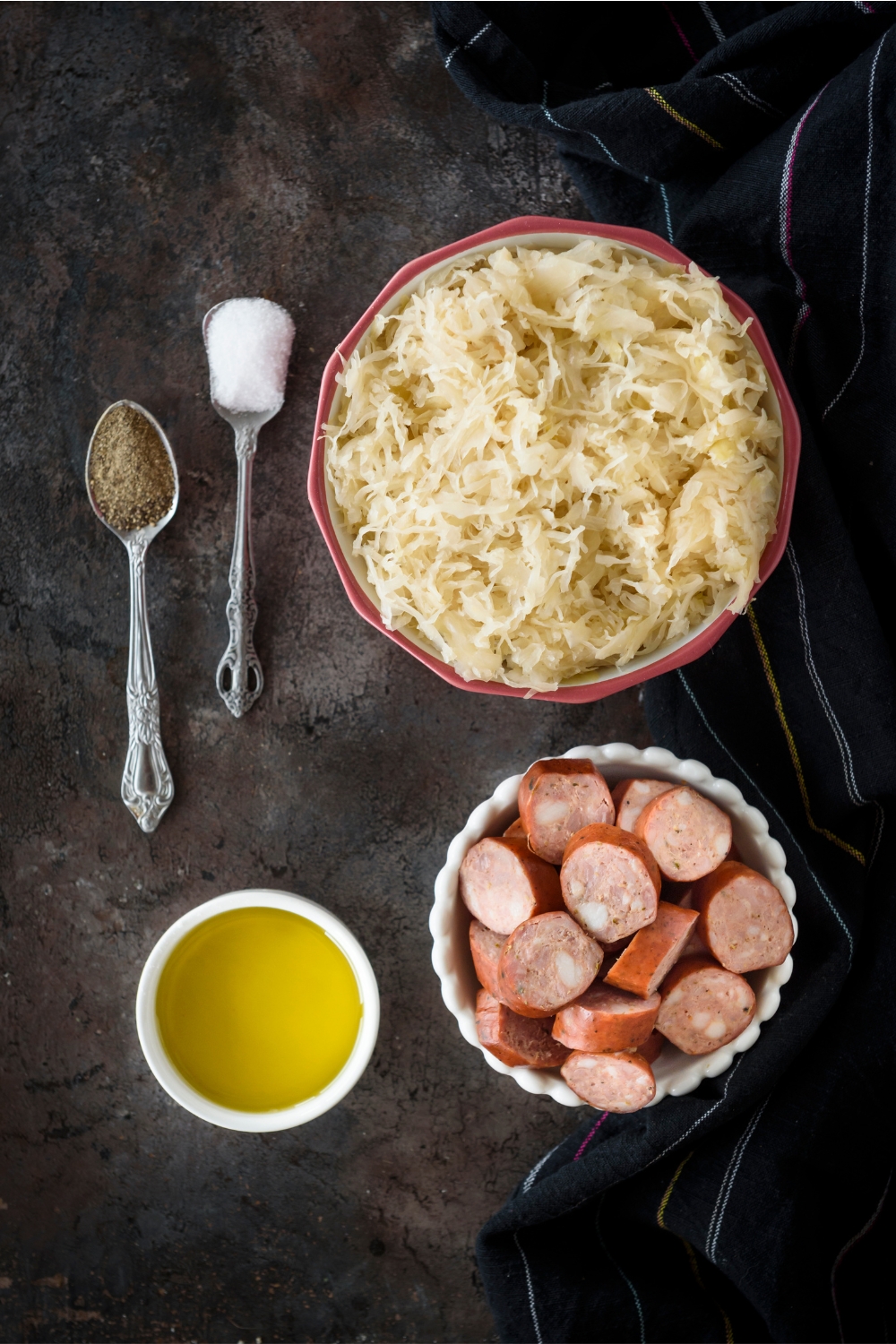 An assortment of ingredients including bowls of sauerkraut, sliced sausages, oil, and two spoonfuls of salt and black pepper.