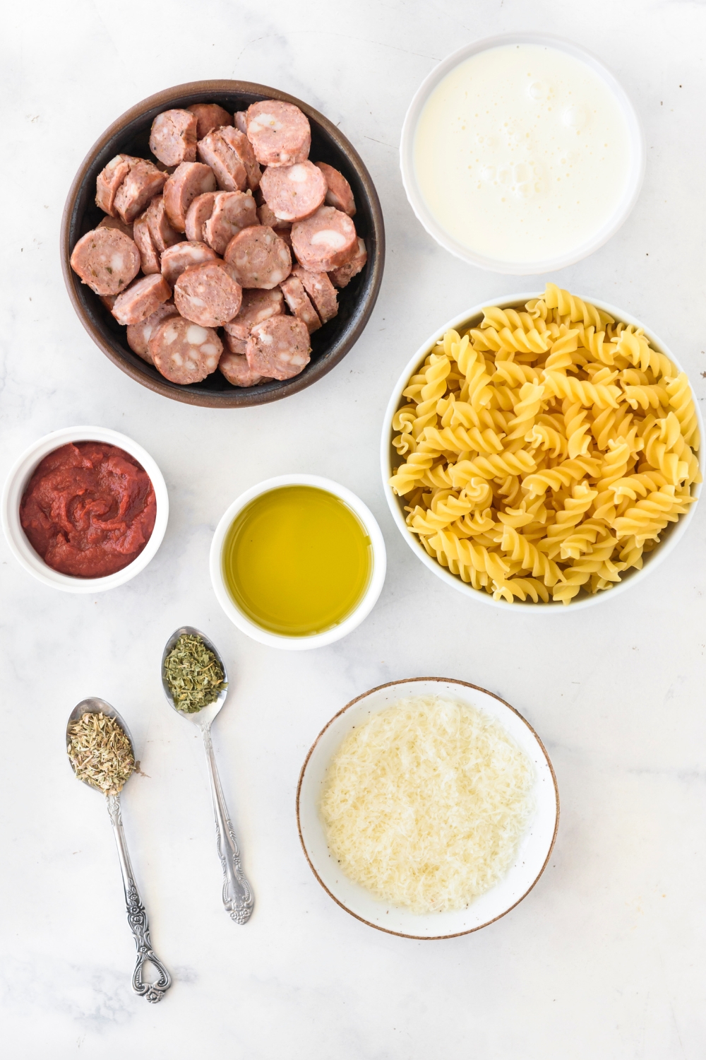 An assortment of ingredients including bowls of sausage slices, dried pasta, tomato paste, cream, cheese, oil, and two spoons full of dried herbs.