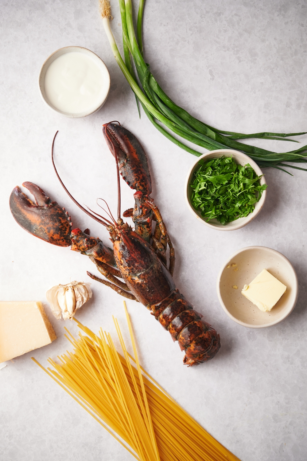 An assortment of ingredients including a lobster, a bunch of scallions, a bunch of dried pasta, a block of parmesan cheese, some garlic cloves, and bowls of cream, butter, and fresh herbs.