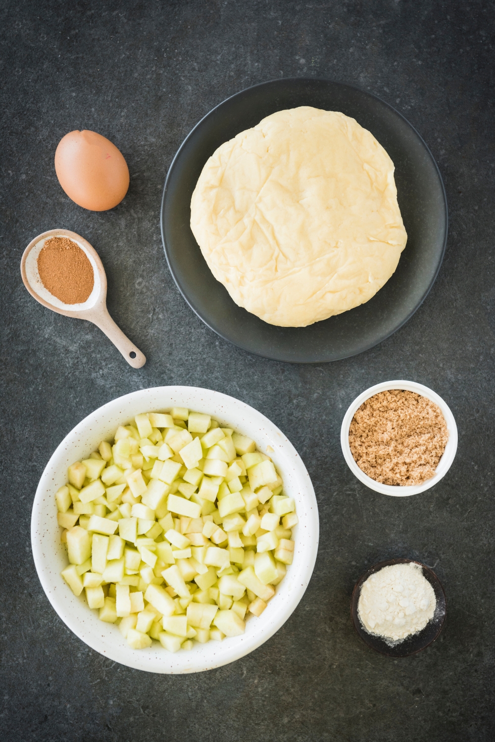 An assortment of ingredients including bowls of diced apples, pie dough, brown sugar, flour, a spoonful of cinnamon, and a single egg, all on a black countertop.