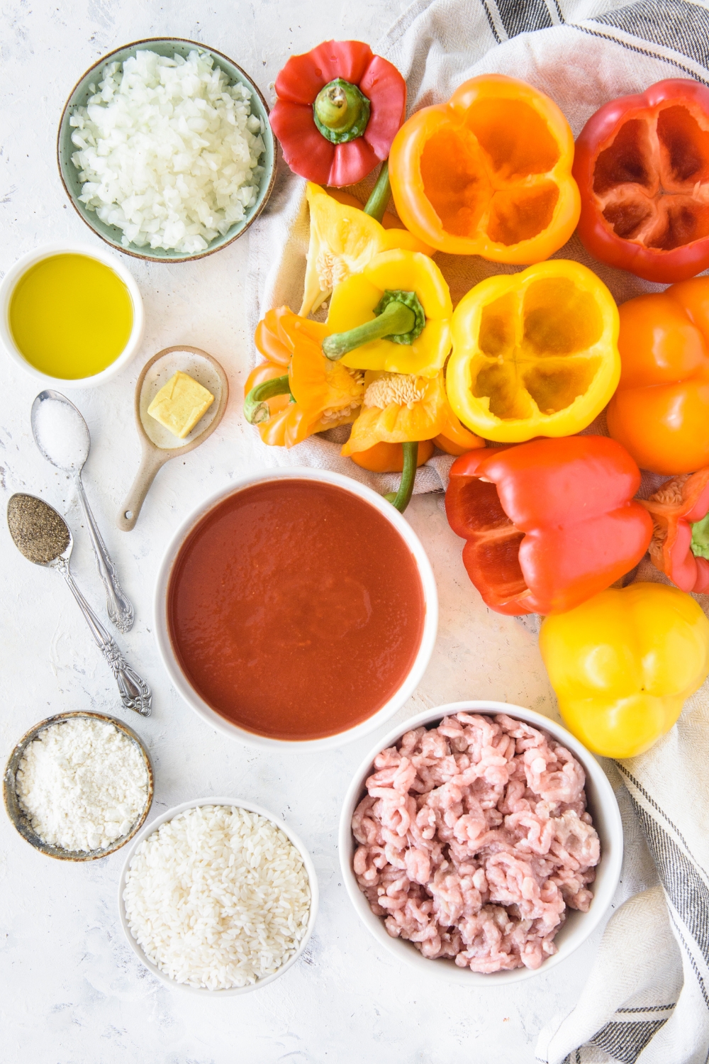 A pile of bell peppers with the seeds and stems removed next to bowls of ingredients including raw ground meat, tomato sauce, flour, oil, diced onion, and butter.