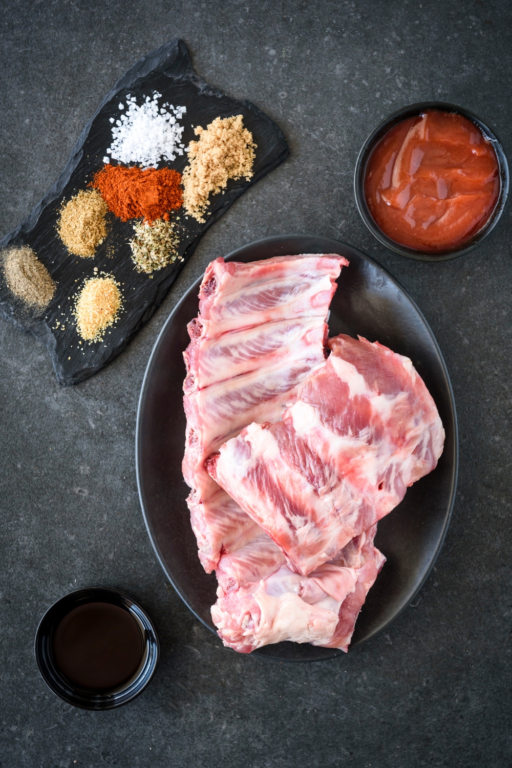 A plate of raw ribs, a bowl of BBQ sauce, a platter with an assortment of seasonings, and a bowl of oil.