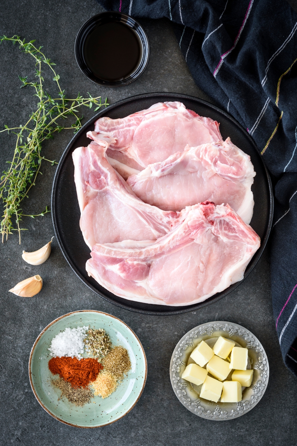 An assortment of ingredients including a plate of raw pork chops, a plate of seasonings, a plate of butter cubes, and a bowl of oil.