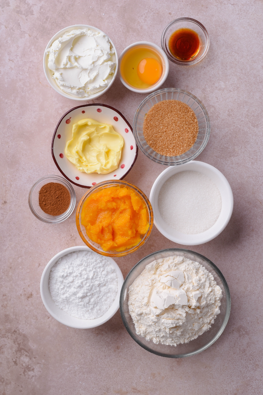 An assortment of ingredients including bowls of pumpkin puree, butter, flour, sugar, vanilla extract, spices, cream cheese, and an unbeaten egg.