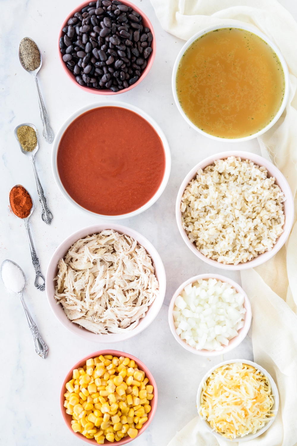 An assortment of ingredients including bowls of black beans, tomato sauce, broth, shredded chicken, rice, corn, shredded cheese, onion, and spoonfuls of spices.