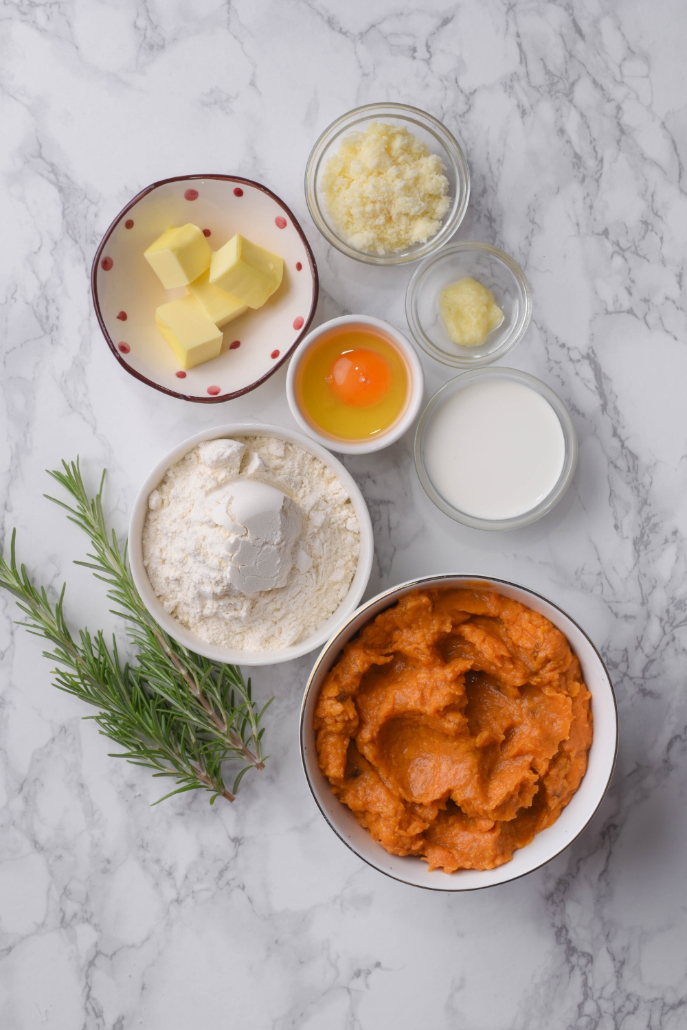 An assortment of ingredients including bowls of mashed sweet potato, an egg, butter cubes, flour, cream, parmesan cheese, and two sprigs of rosemary on a grey counter.