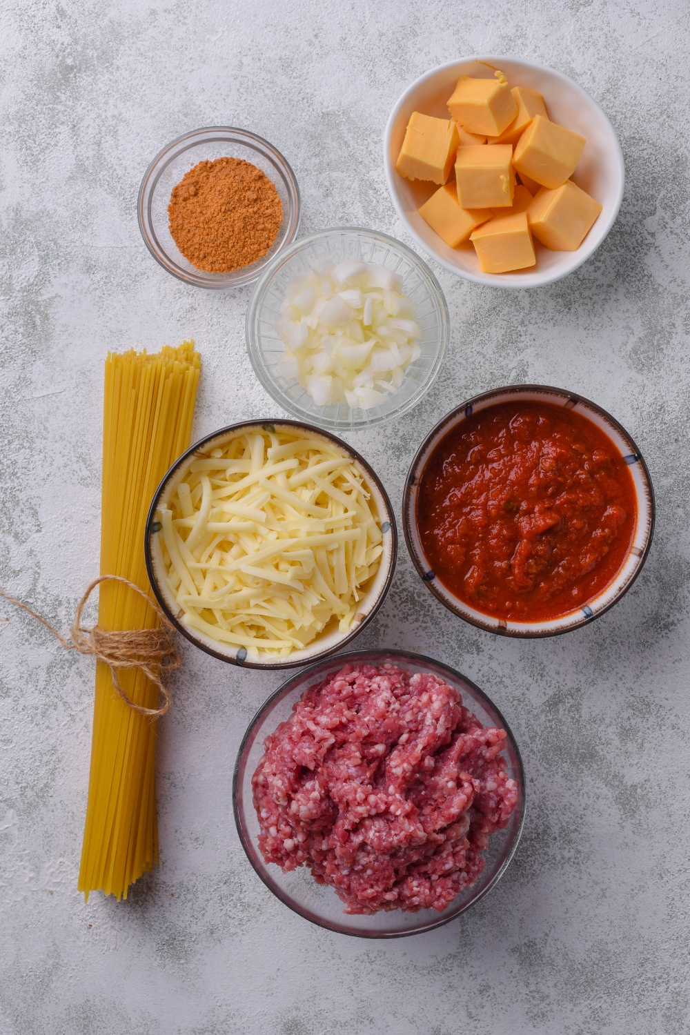 An assortment of ingredients including a bundle of dried spaghetti noodles and bowls of raw ground beef, cubes of Velveeta cheese, shredded cheese, salsa, diced onion, and spices, all on a grey countertop.