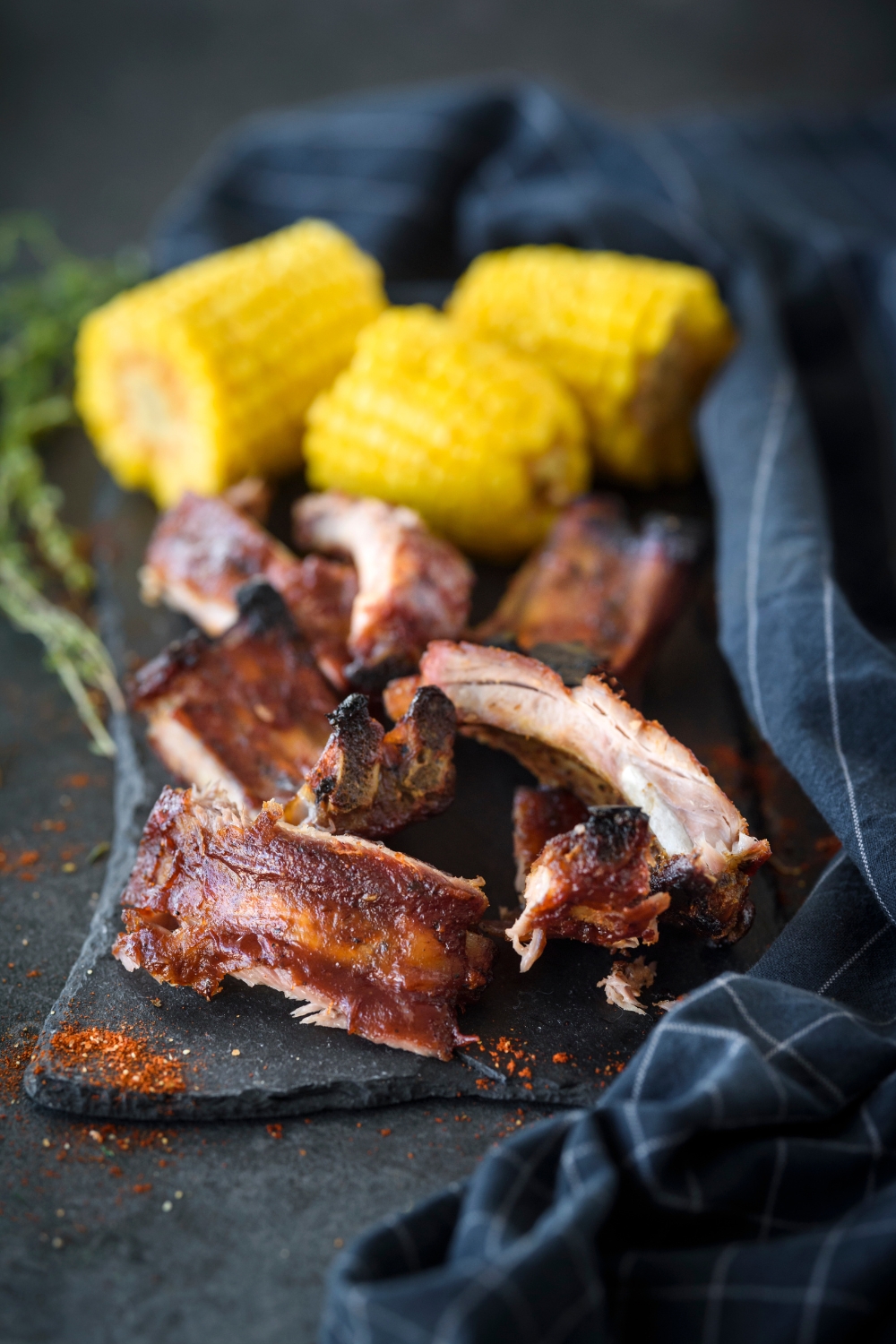 A pile of ribs covered in BBQ sauce on a black platter with three sliced corn cobs.