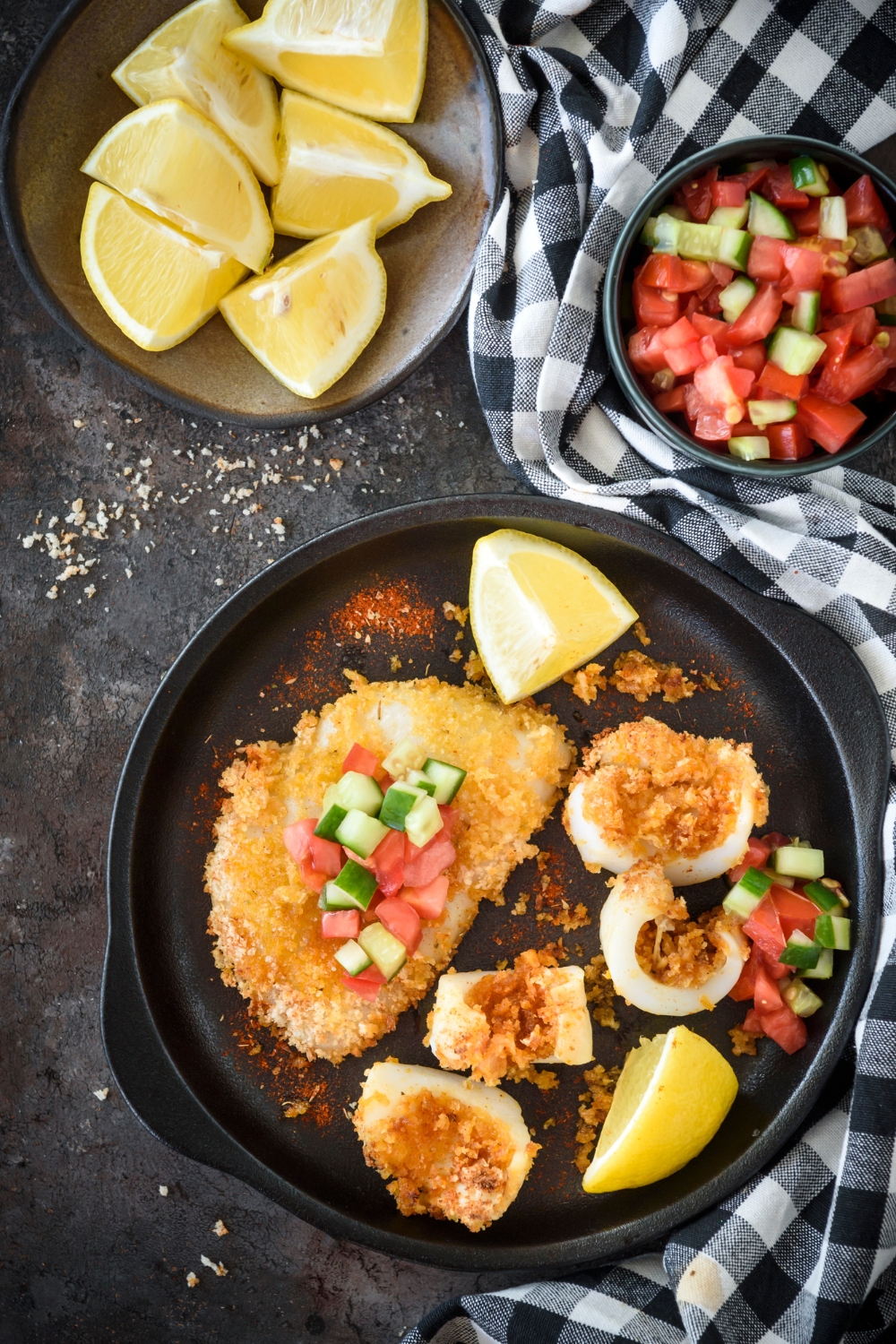 A breaded calamari steak topped with cucumber and diced tomato on a plate with a lemon wedge and chunks of sliced calamari.