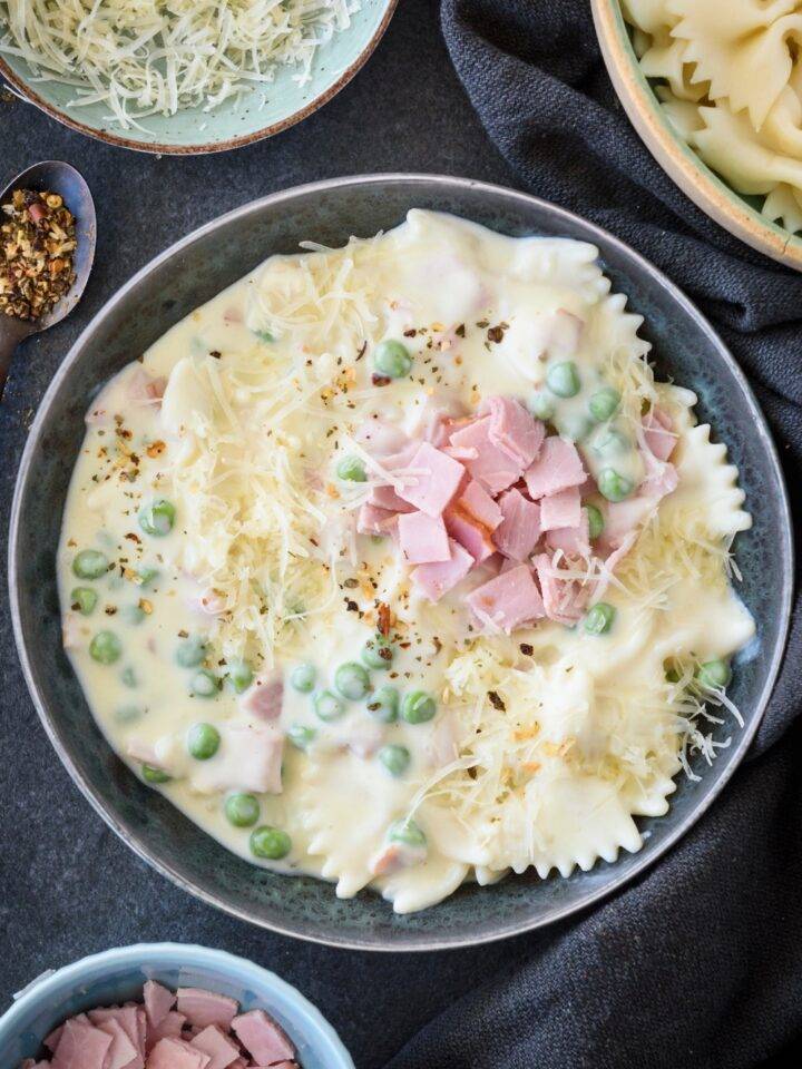 A bowl filled with pasta, ham, peas, and shredded cheese covered in cream sauce.