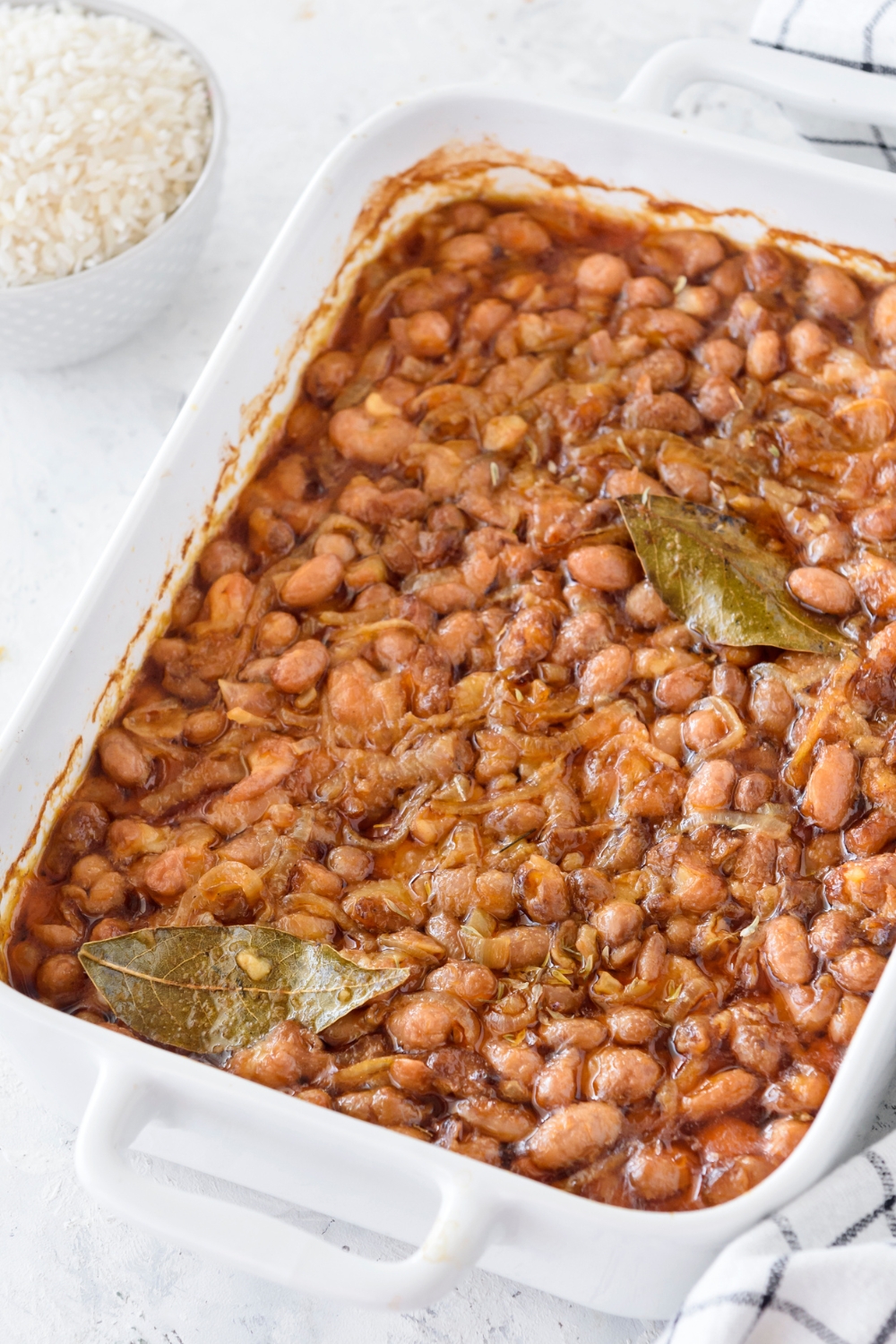 A baking dish filled with cooked pinto beans mixed with sautéed onions and two bay leaves.
