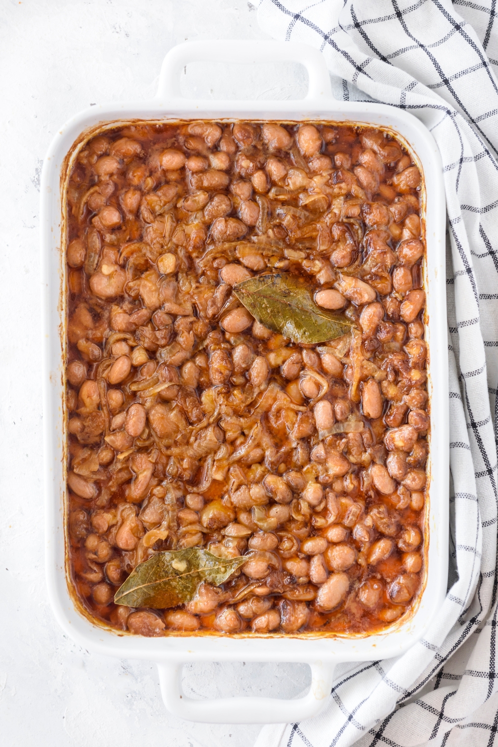 A baking dish filled with cooked pinto beans mixed with sautéed onions and two bay leaves.