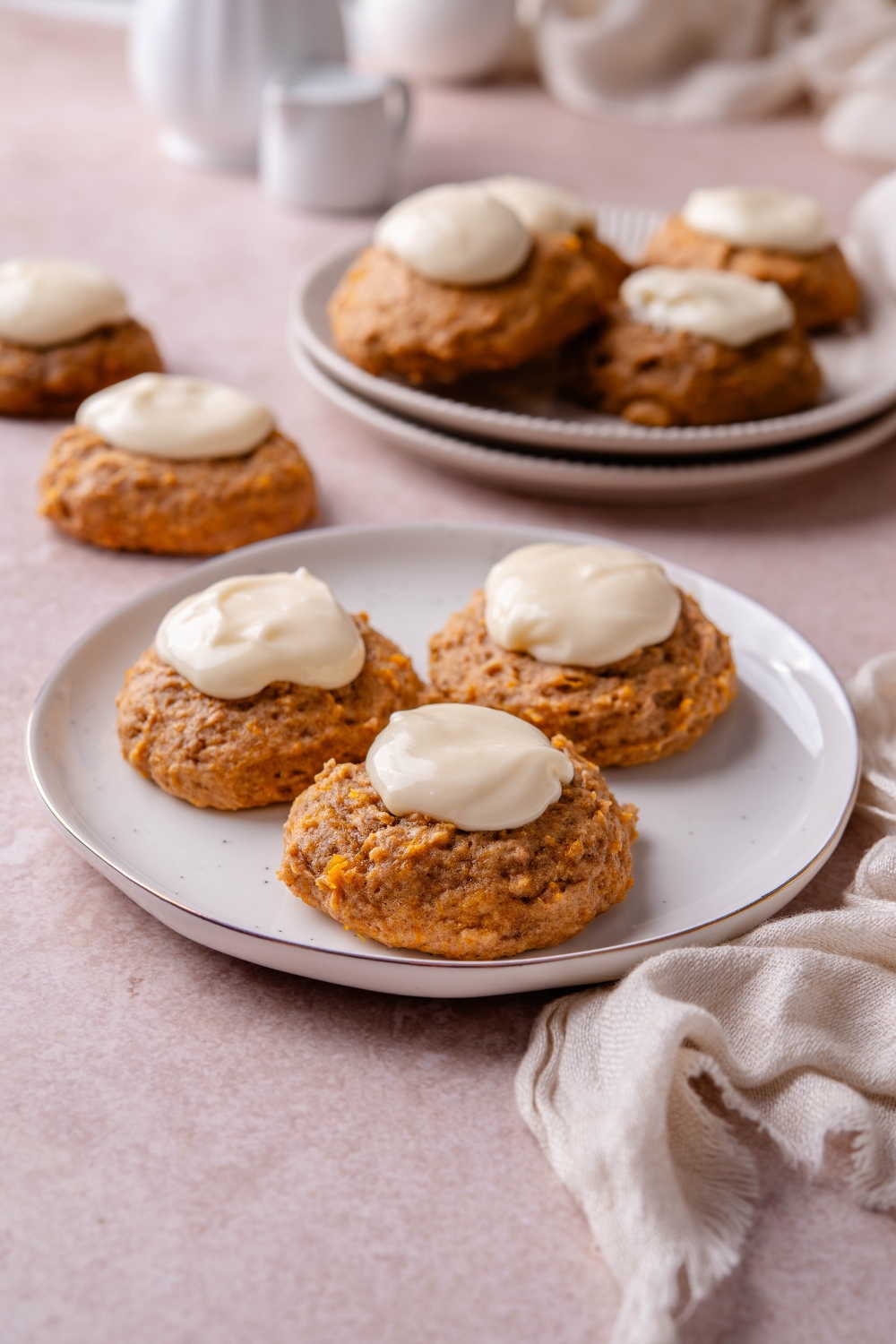 Three pumpkin cookies topped with frosting on a white plate with more cookies on a plate and on the counter in the background.