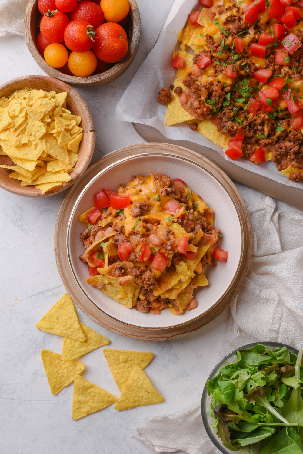 A bowl of beef nachos topped with melted cheese and diced tomatoes. Next to the bowl is a baking sheet filled with more beef nachos.