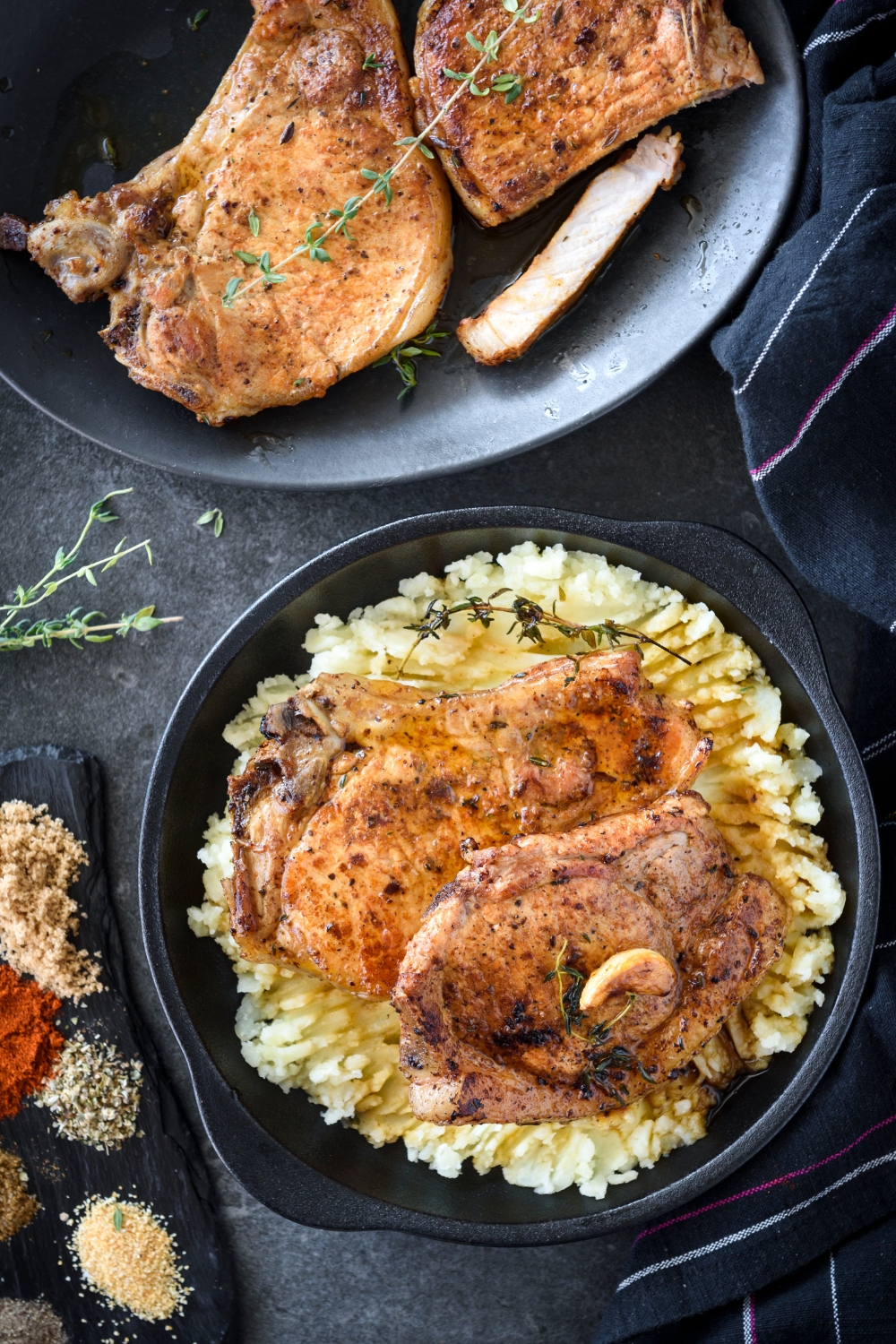 A plate with two cooked and seasoned pork chops on a bed of mashed potatoes with garlic cloves and herbs on the pork chops. Next to the plate is a second plate with two cooked pork chops.