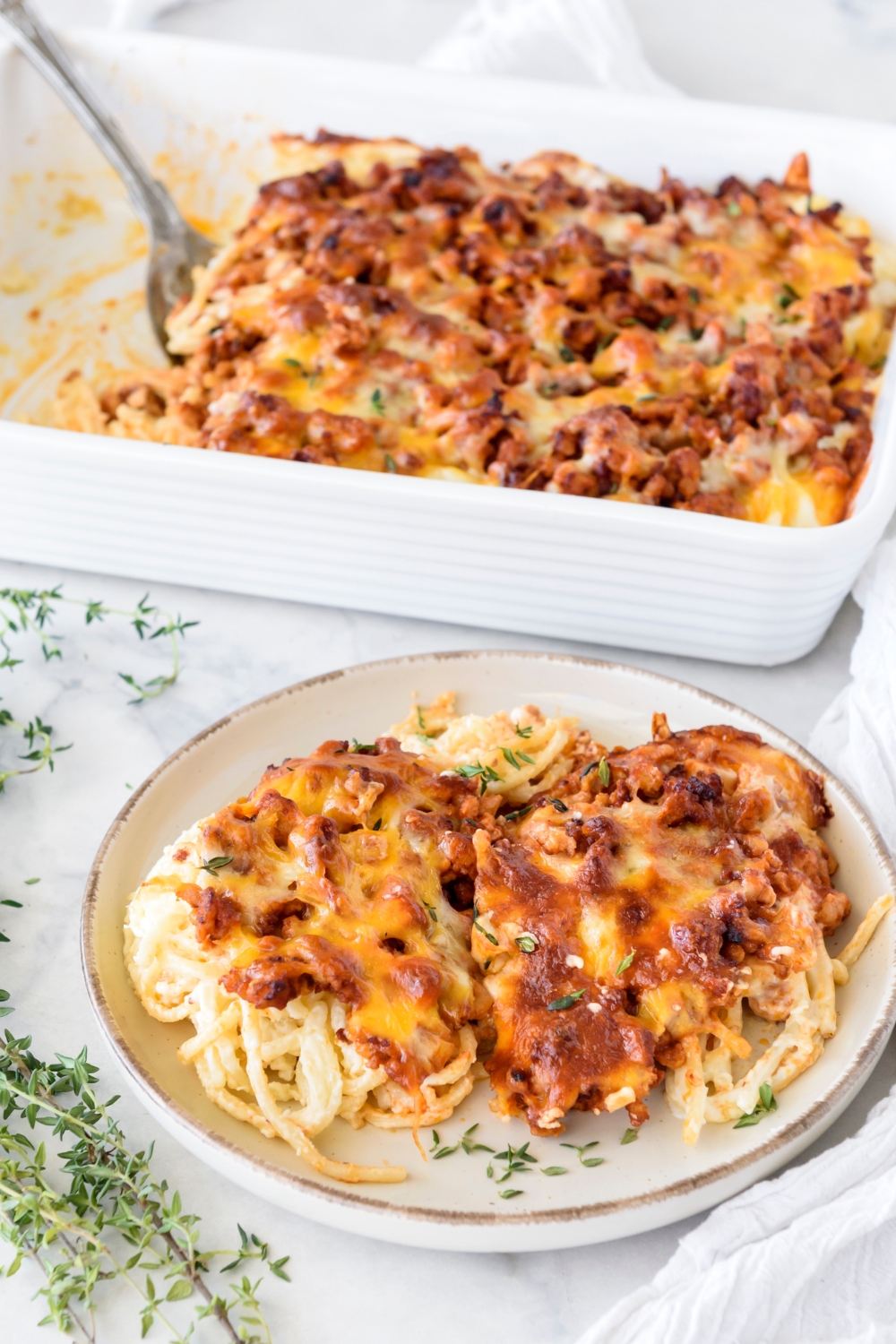 A bowl of baked spaghetti covered in melted cheese with chunks of sausage. Next to the plate is a baking dish filled with baked spaghetti.