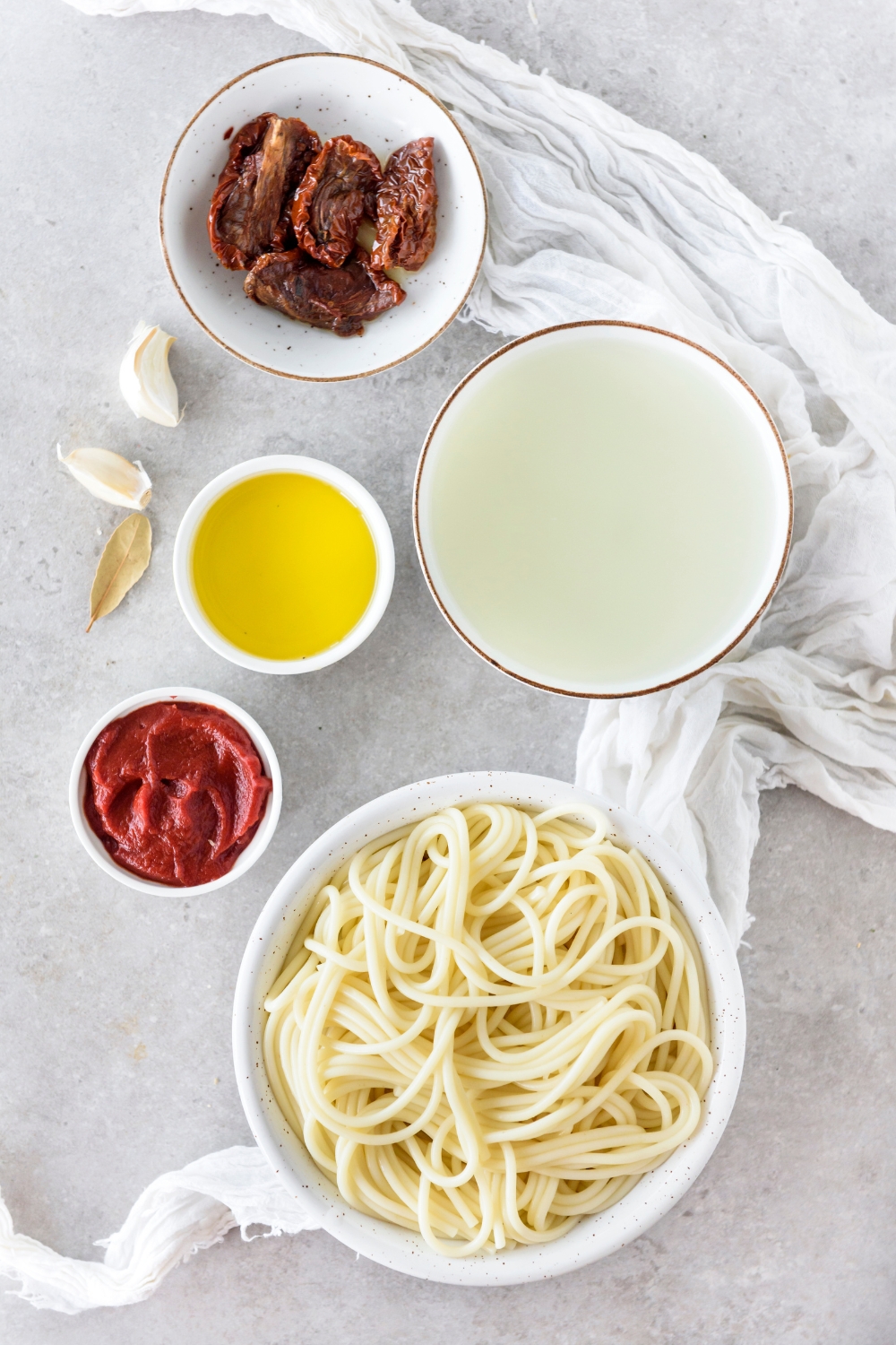 An assortment of ingredients including bowls of spaghetti noodles, sundried tomatoes, tomato paste, oil, and water.