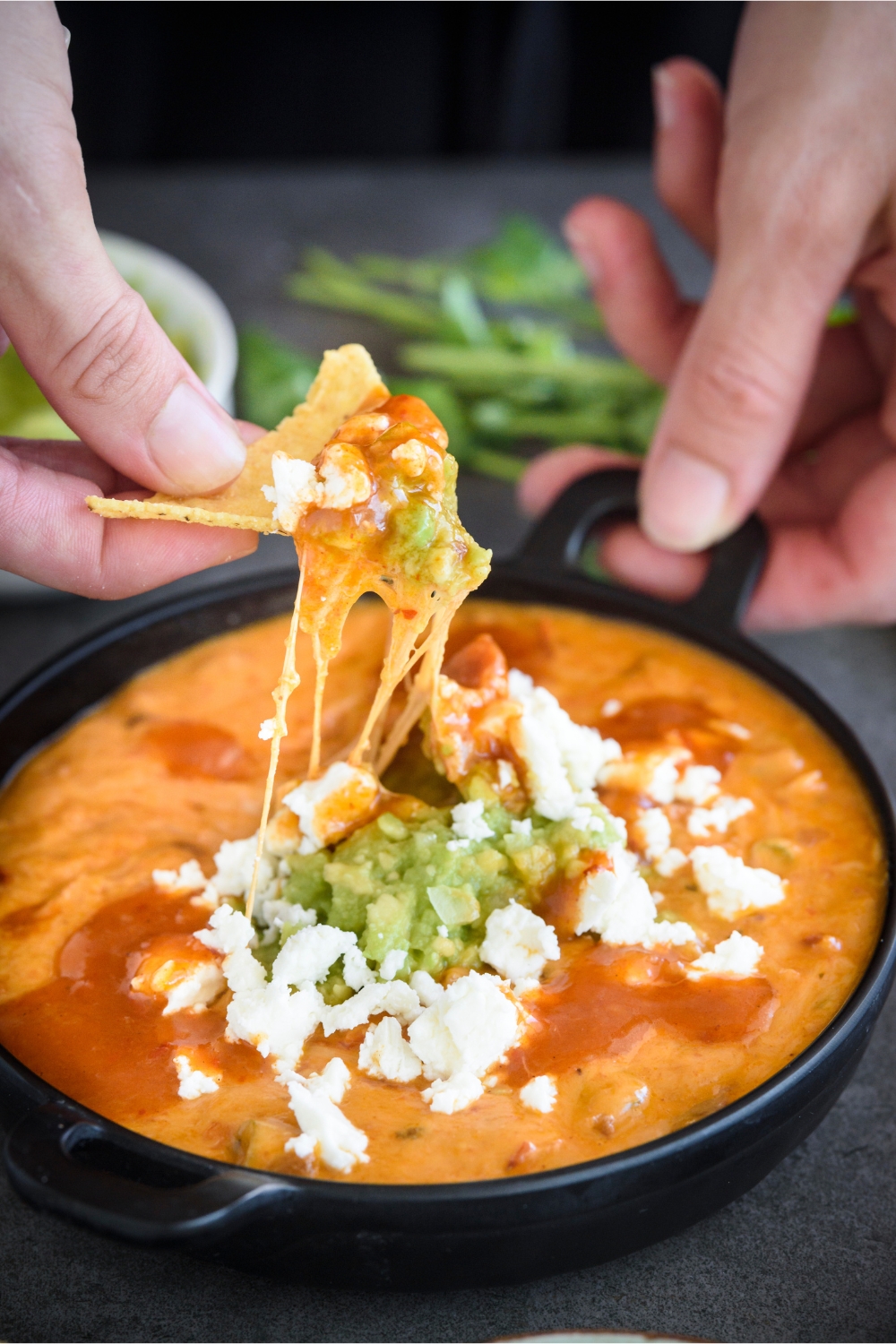 A tortilla chip with a heaping scoop of queso being held over the serving bowl.