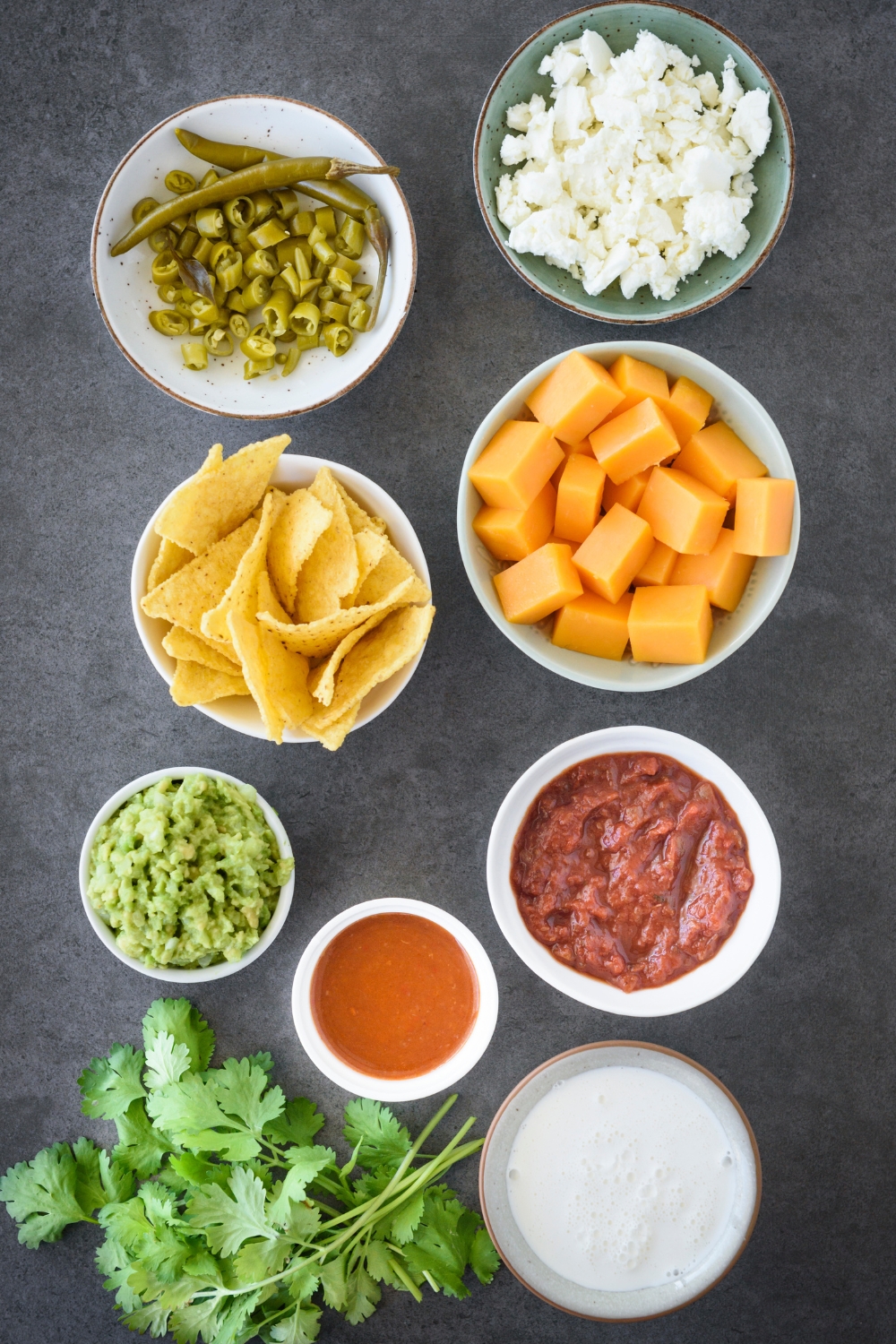 A countertop with cheese, tortilla chips, green chiles, salsa, hot sauce, guacamole, heavy cream, and cilantro in separate dishes.