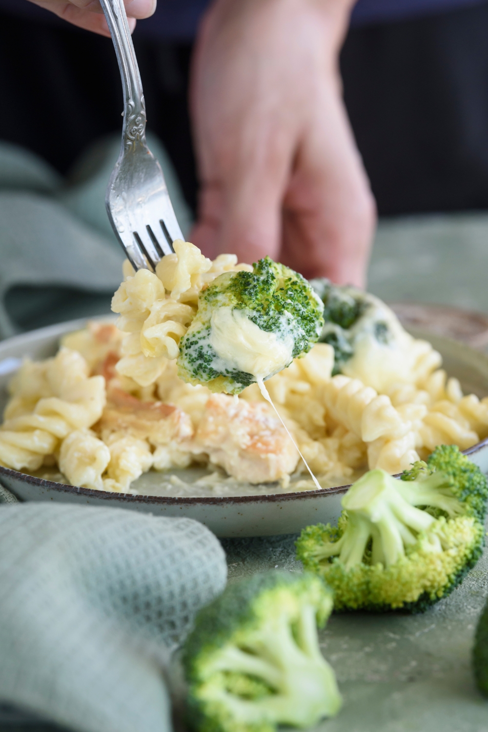 A fork holding a bite of pasta in cream sauce with a piece of broccoli at the end of the fork. Cheese is stringing from the broccoli to the plate of pasta.