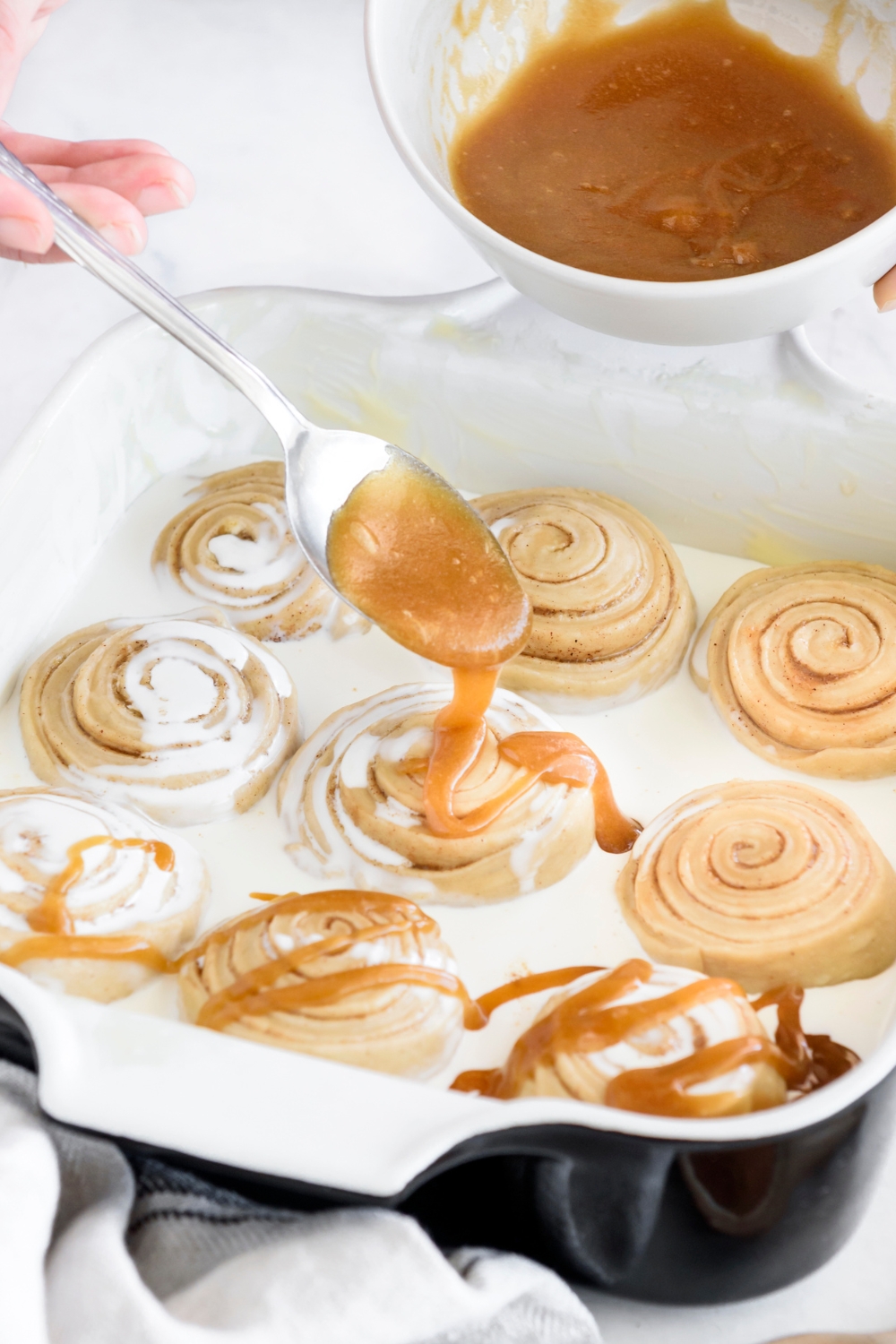 A spoon drizzling a brown sugar glaze onto cinnamon rolls in a baking dish that are covered with cream.