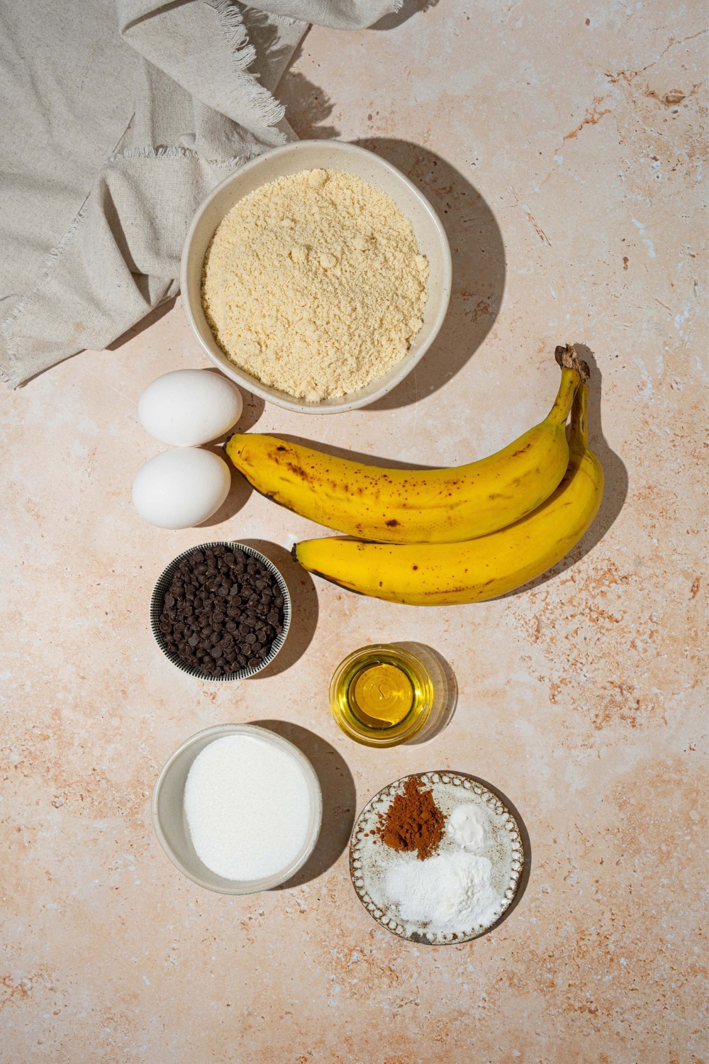 A bowl of almond flour, two eggs, two bananas, bowl, chocolate chips, a bowl of cinnamon salt and baking powder, and a small cup of vanilla extract.