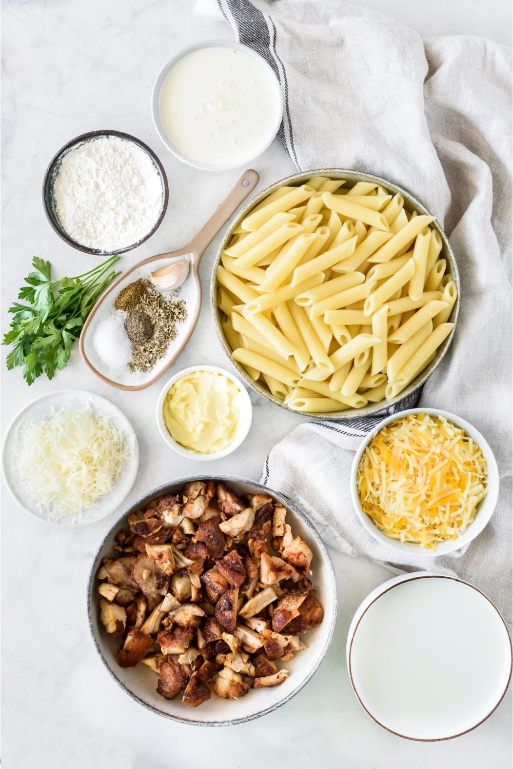 A countertop with multiple bowls containing cooked pasta, seasonings, chicken, cheese, flour, and heavy cream.