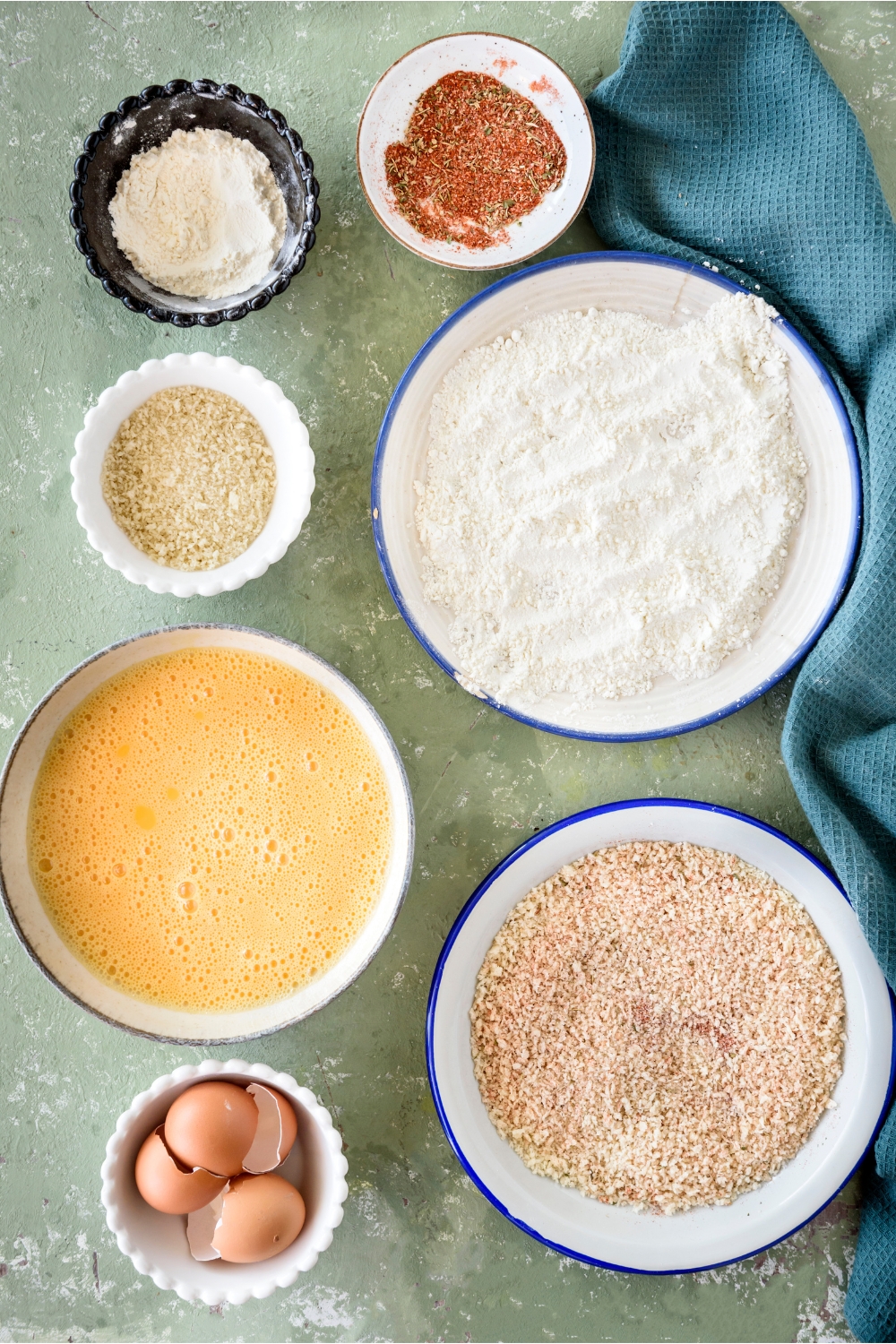 An assortment of ingredients including bowls of flour, seasoning, beaten eggs, egg shells, and breadcrumbs.