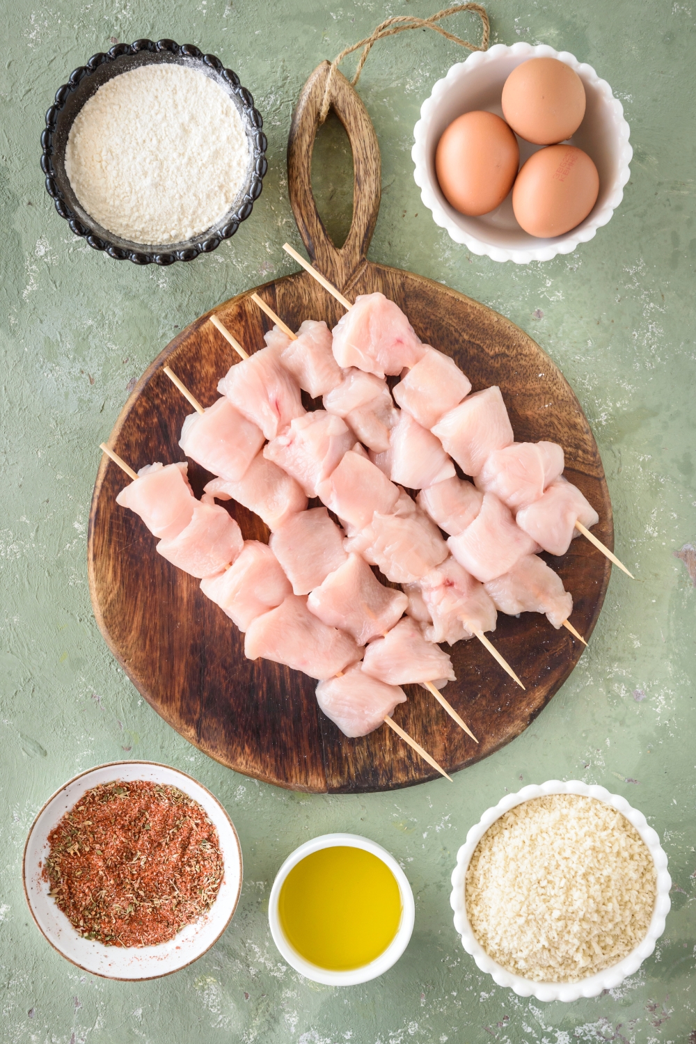 A wooden board filled with raw cubed chicken on skewers, surrounded by bowls of ingredients including eggs, oil, breadcrumbs, and seasoning.
