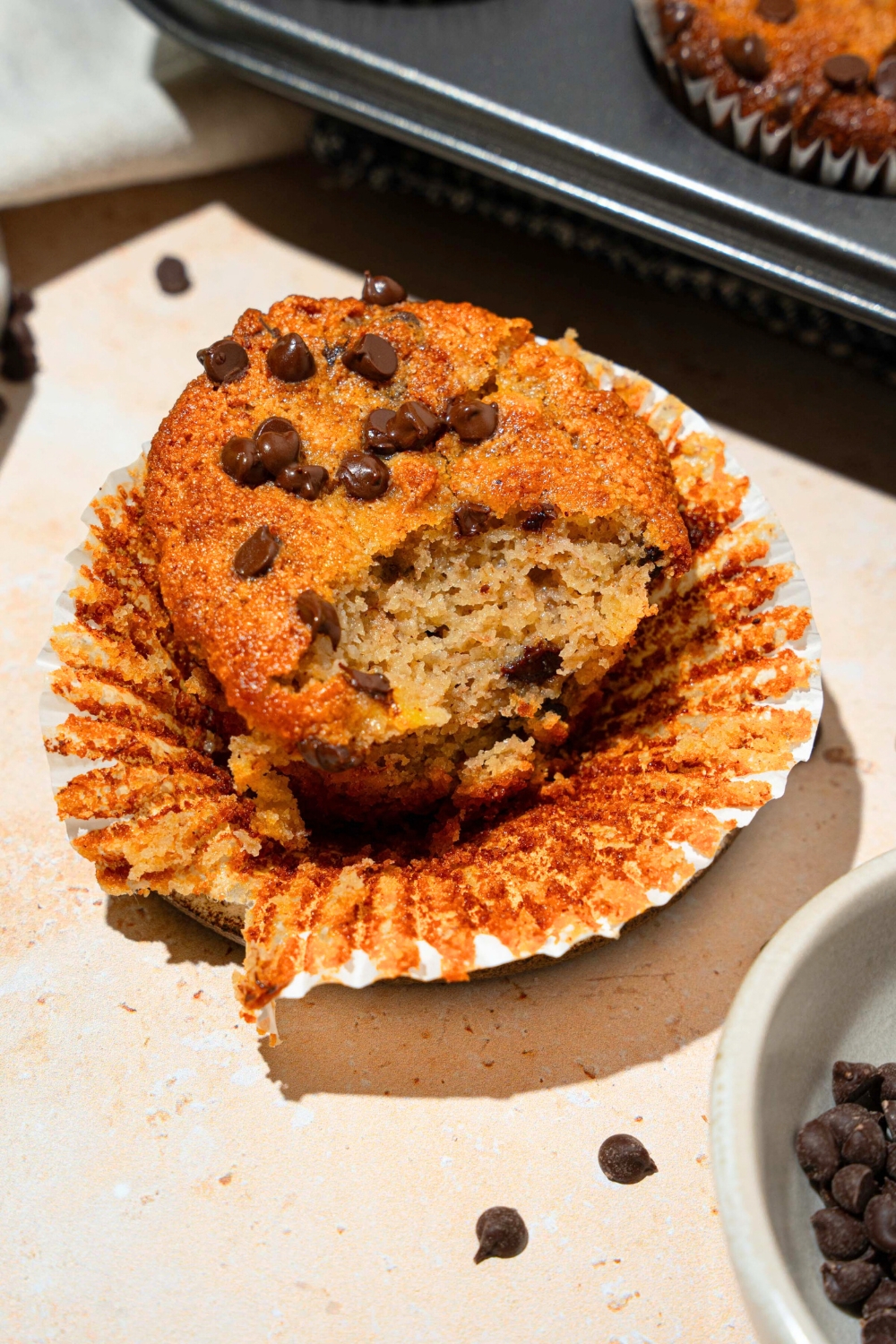 A chocolate chip almond flour, banana muffin with a wrapper opening up away from it and a bite out of the front of it.