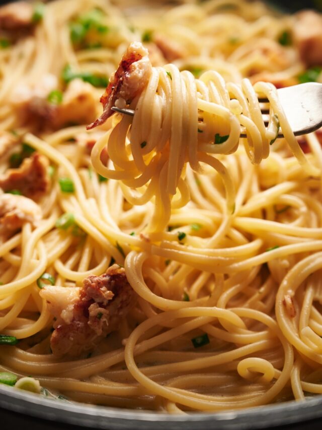 A fork holding a bite of cooked spaghetti with a piece of lobster meat at the end of the fork. The fork is held above a skillet filled with spaghetti.