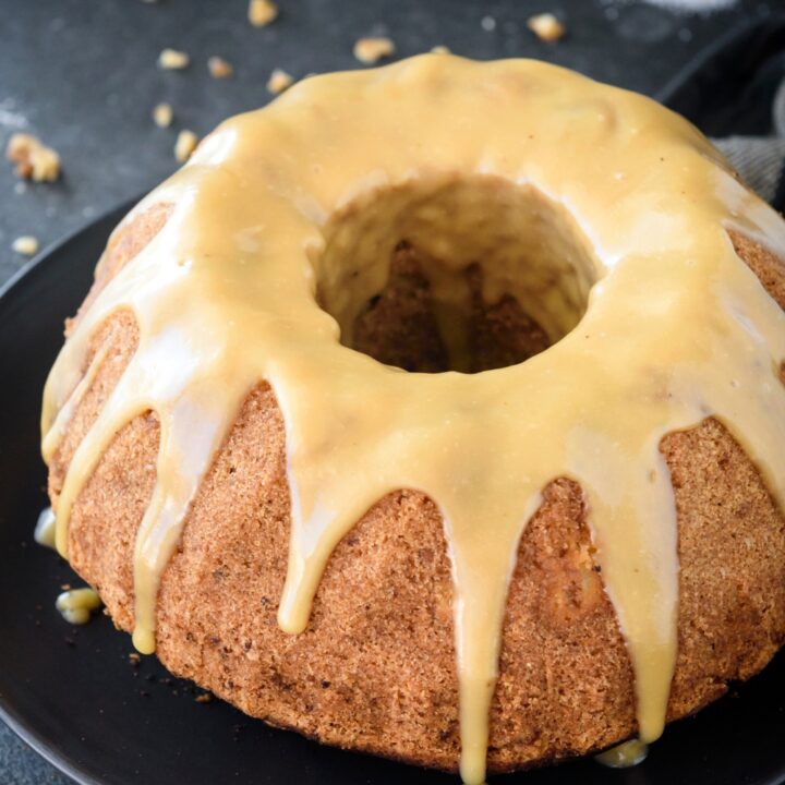 An apple bundt cake with a caramel glaze on top. The cake is on a black plate on a black counter.