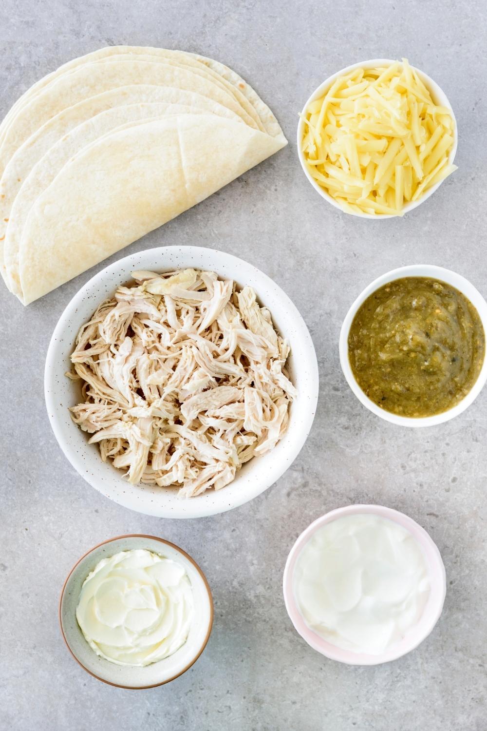 A countertop with multiple bowls with green enchilada sauce, shredded cheese, shredded chicken, sour cream, and cream cheese.