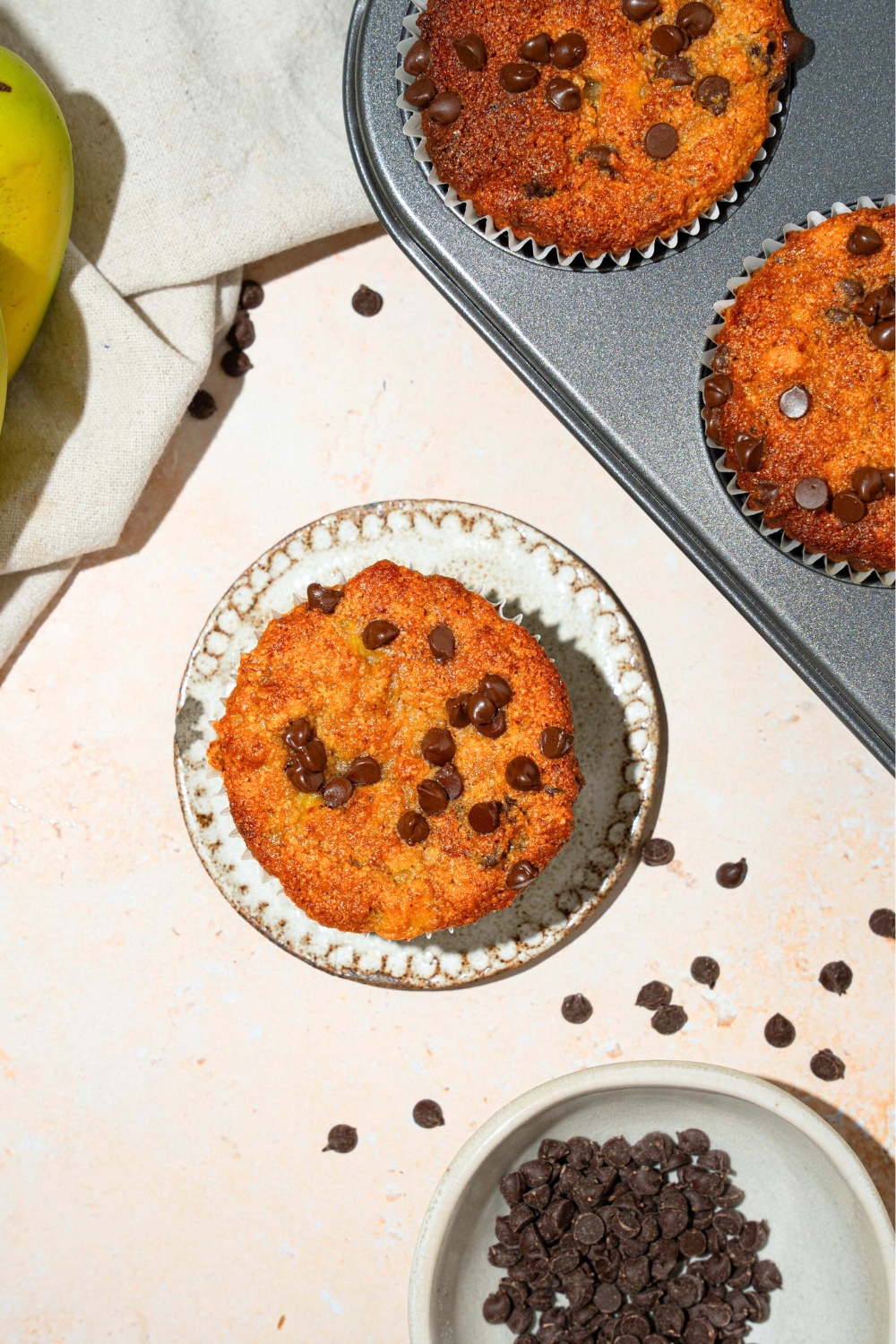 An overhead picture of an almond flower chocolate chip banana muffin on a white plate with a tin of the muffins behind it.