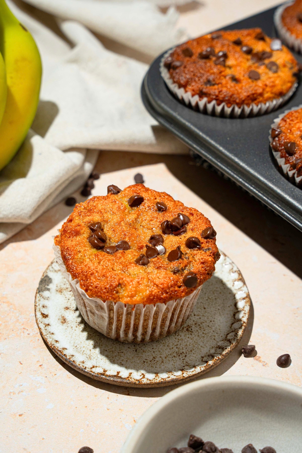 A chocolate chip almond flour, banana muffin on a white plate with part of the tin of the muffins behind it.