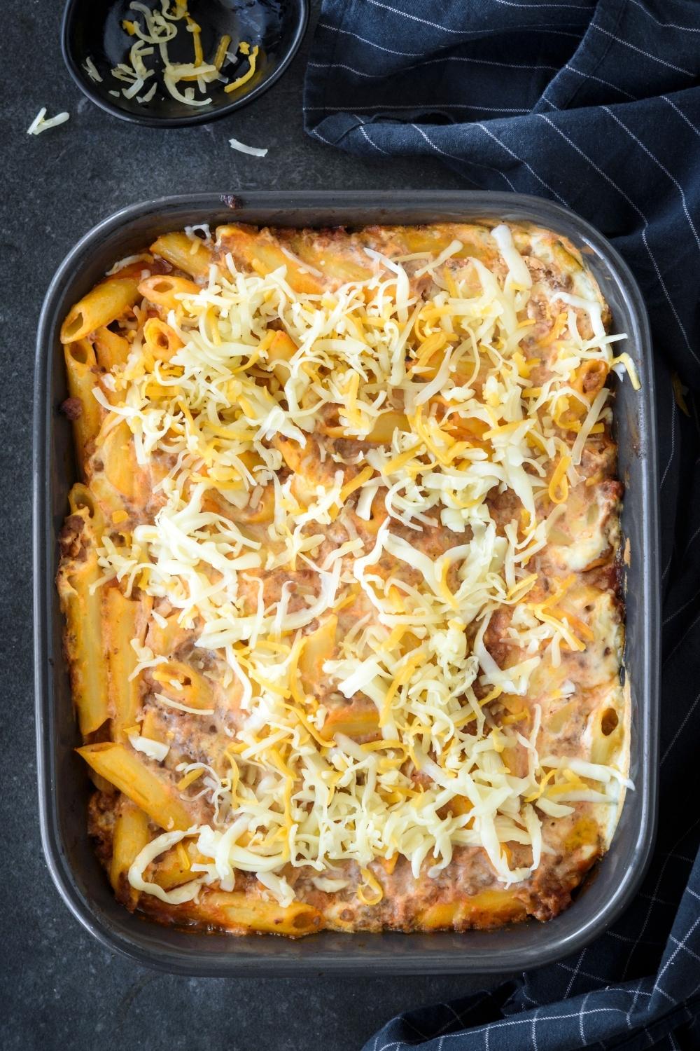 A casserole dish with baked casserole being topped with shredded cheese.