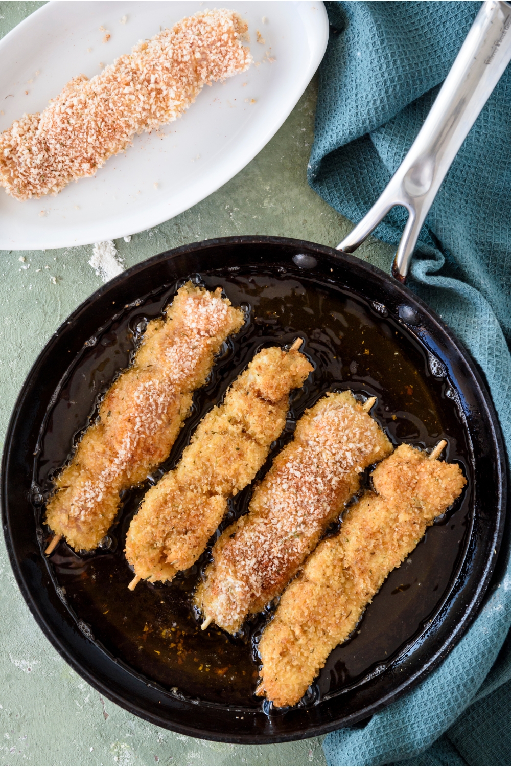 A pan filled with oil and four breaded chicken skewers frying in it.