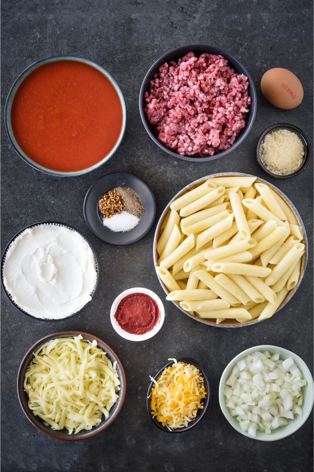 A countertop with tomato sauce, beef, and egg, parmesan cheese, seasoning, ricotta cheese, pasta noodles, tomato paste, shredded cheeses, and minced onion in separate bowls.