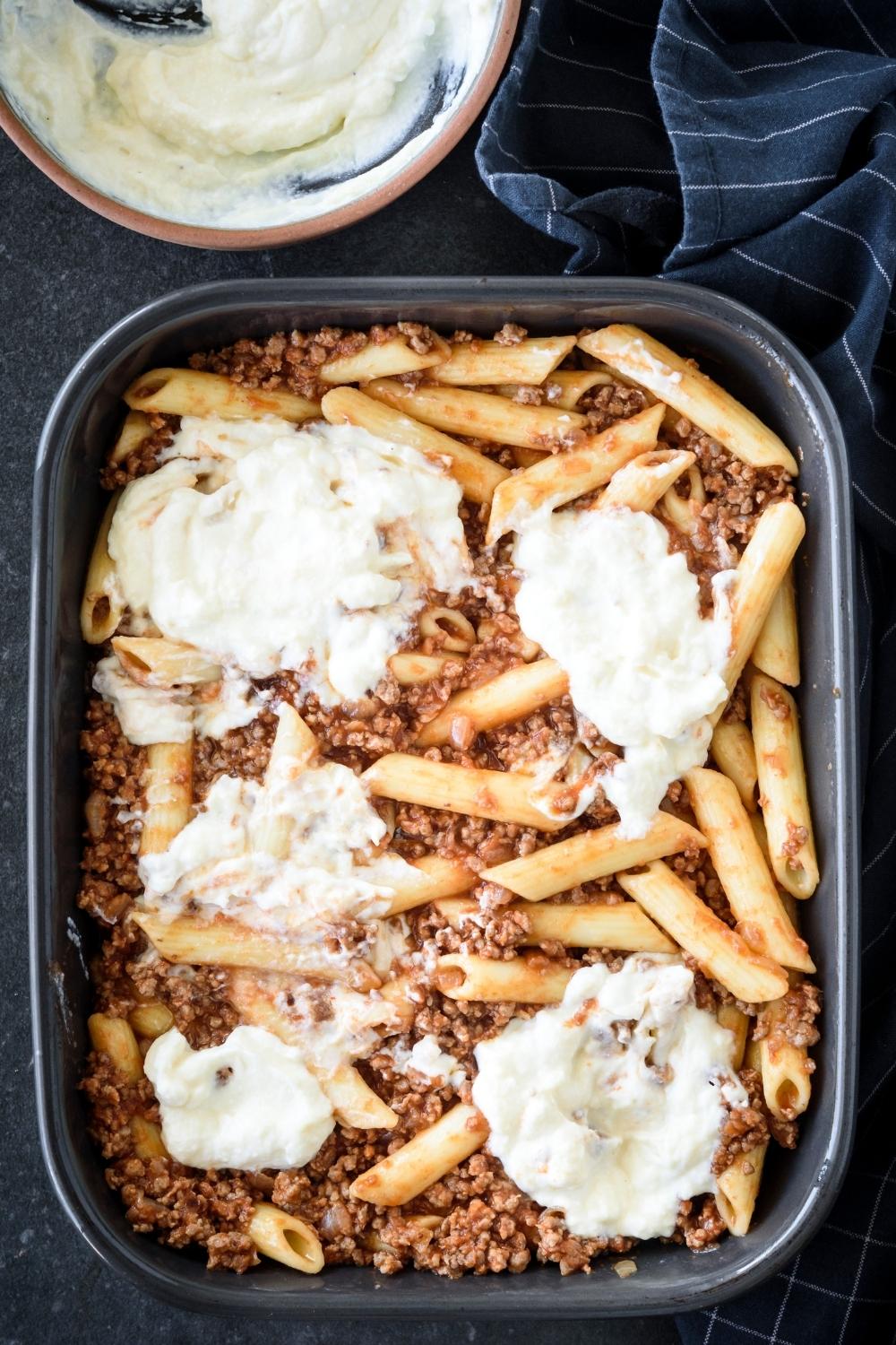 A casserole dish with the cheese mixture being added to the pasta beef mixture.