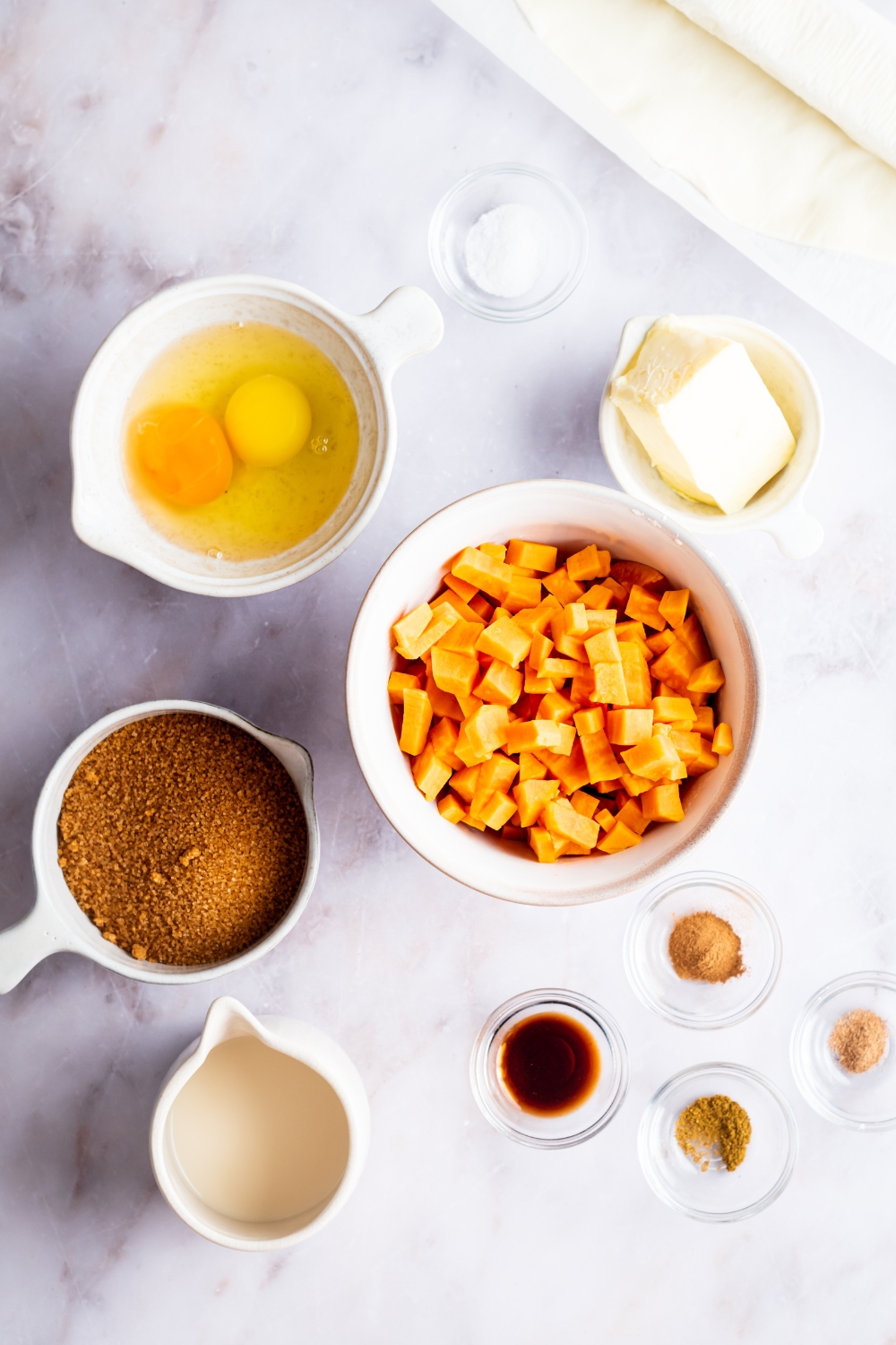 A bowl of two eggs, a cup of butter, a bowl of sweet potatoes, a bowl of brown sugar, a pitcher of milk, and small bowls of spices.