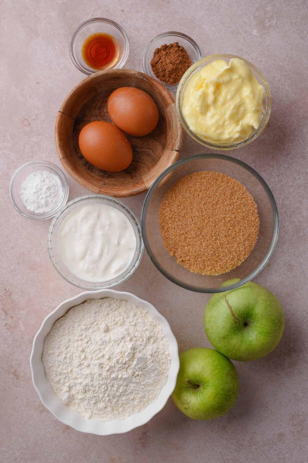 A countertop with various bowls containing butter, eggs, vanilla, apple pie spice, brown sugar, sour cream, baking powder, flour, and apples.