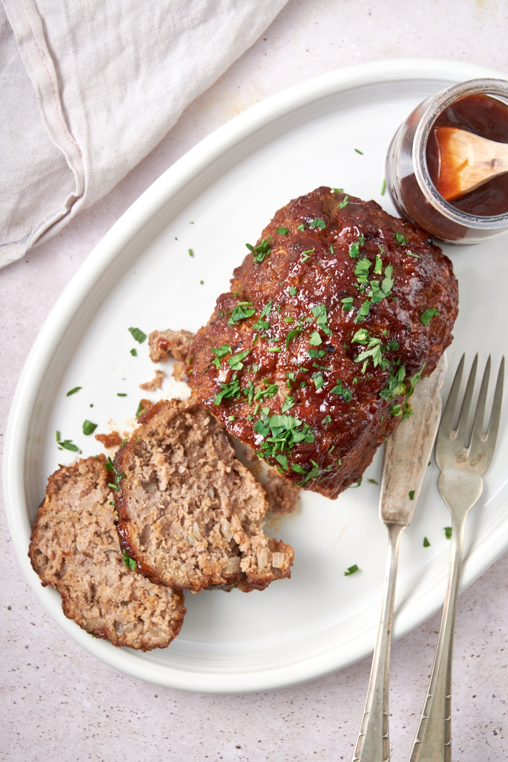 A cooked and sliced barbecue meatloaf garnished with parsley on a white serving tray. A serving knife and fork are nearby with a jar of barbecue sauce.